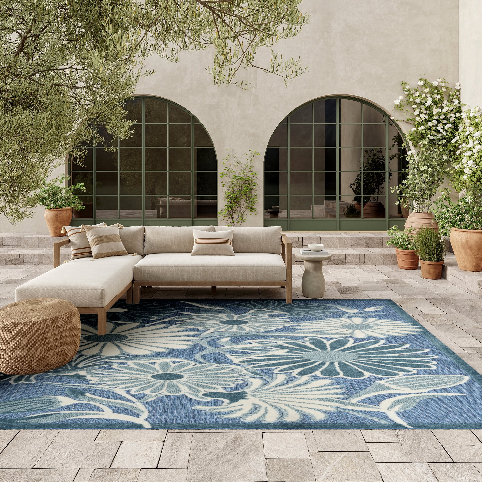 Outdoor beige sectional on a wooden frame, blue floral rug, arched green-framed windows, potted plants.
