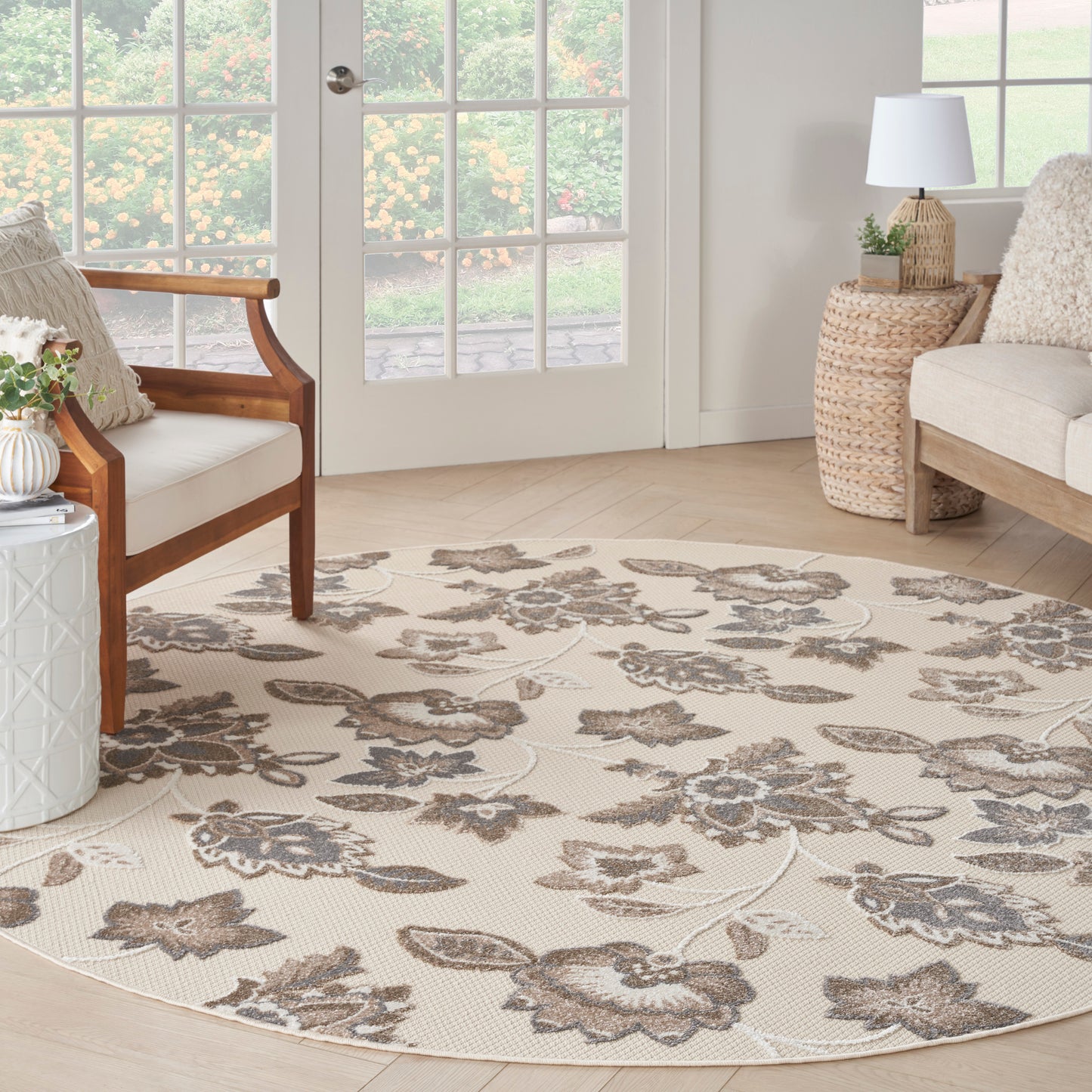 Round beige floral area rug with gray-brown flowers in a cozy neutral living room with wooden chairs and glass doors.