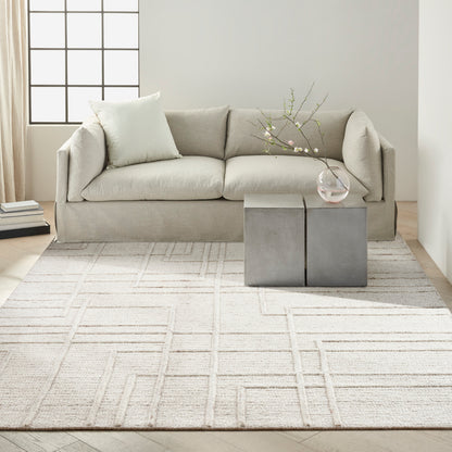 Neutral beige sofa with light cushions on a pale geometric rug; concrete cube side tables with a vase of flowering branches.