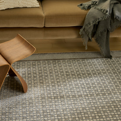 Curved plywood mid-century chair on a gray-beige geometric rug, beside a tan sofa with a fringed gray throw.
