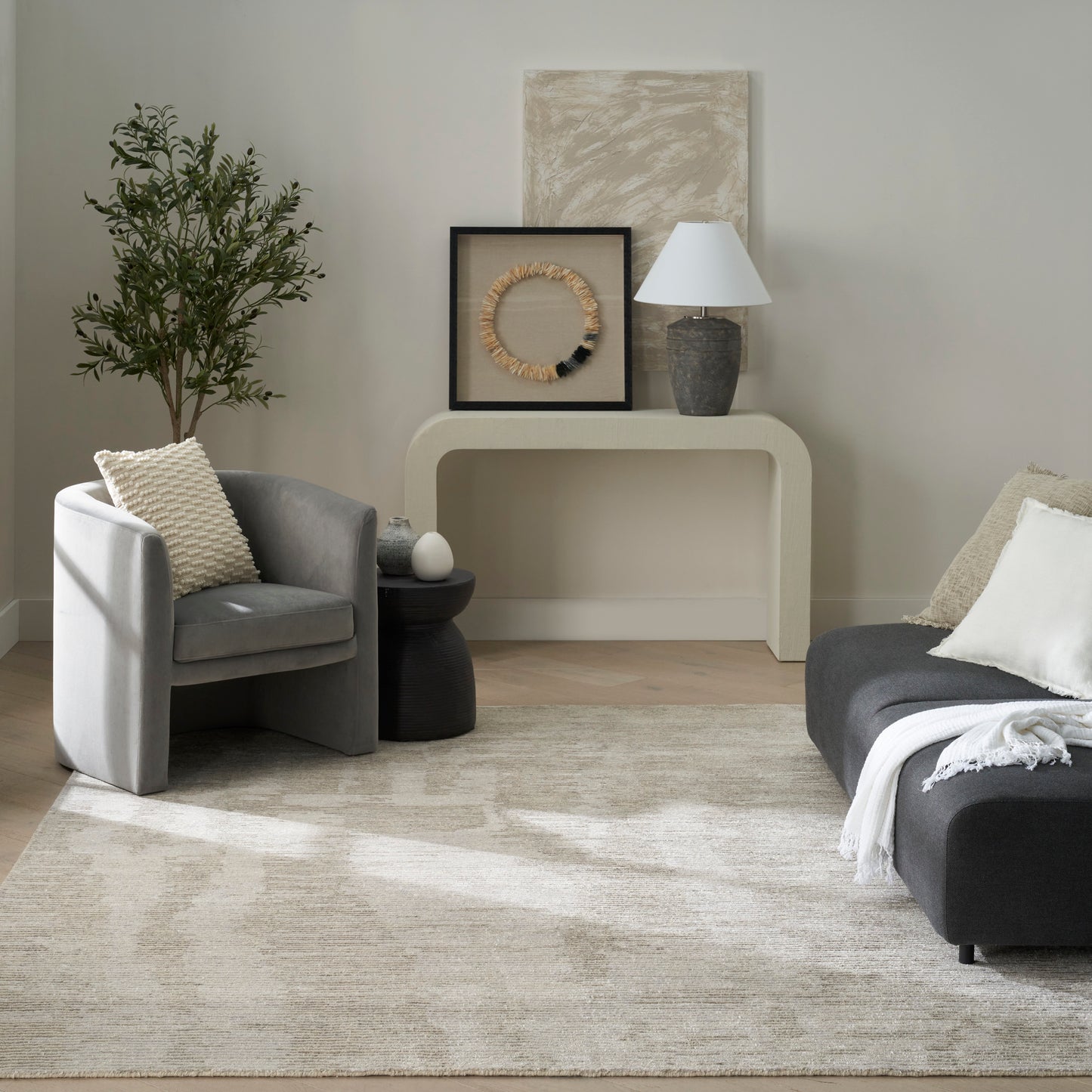 Neutral living room with gray armchair, dark sofa, beige rug, curved cream console table, wicker wall art, lamp, and plant.