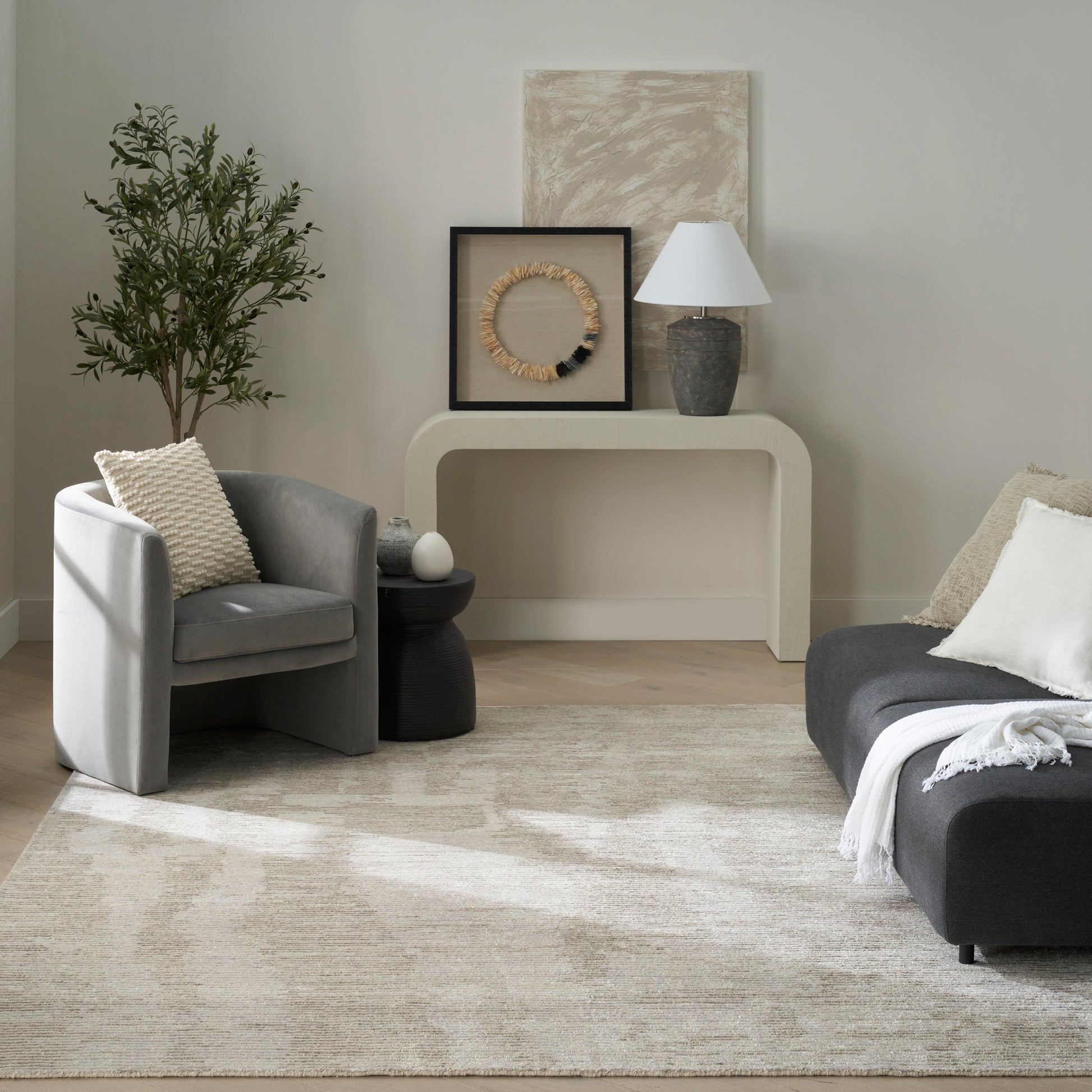 Neutral living room with gray armchair, dark sofa, beige rug, curved cream console table, wicker wall art, lamp, and plant.