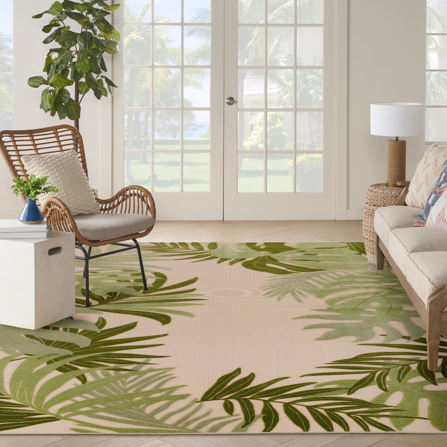 Living room with beige rug featuring green tropical leaves, wicker chair, neutral sofa, potted plant, and glass French doors.