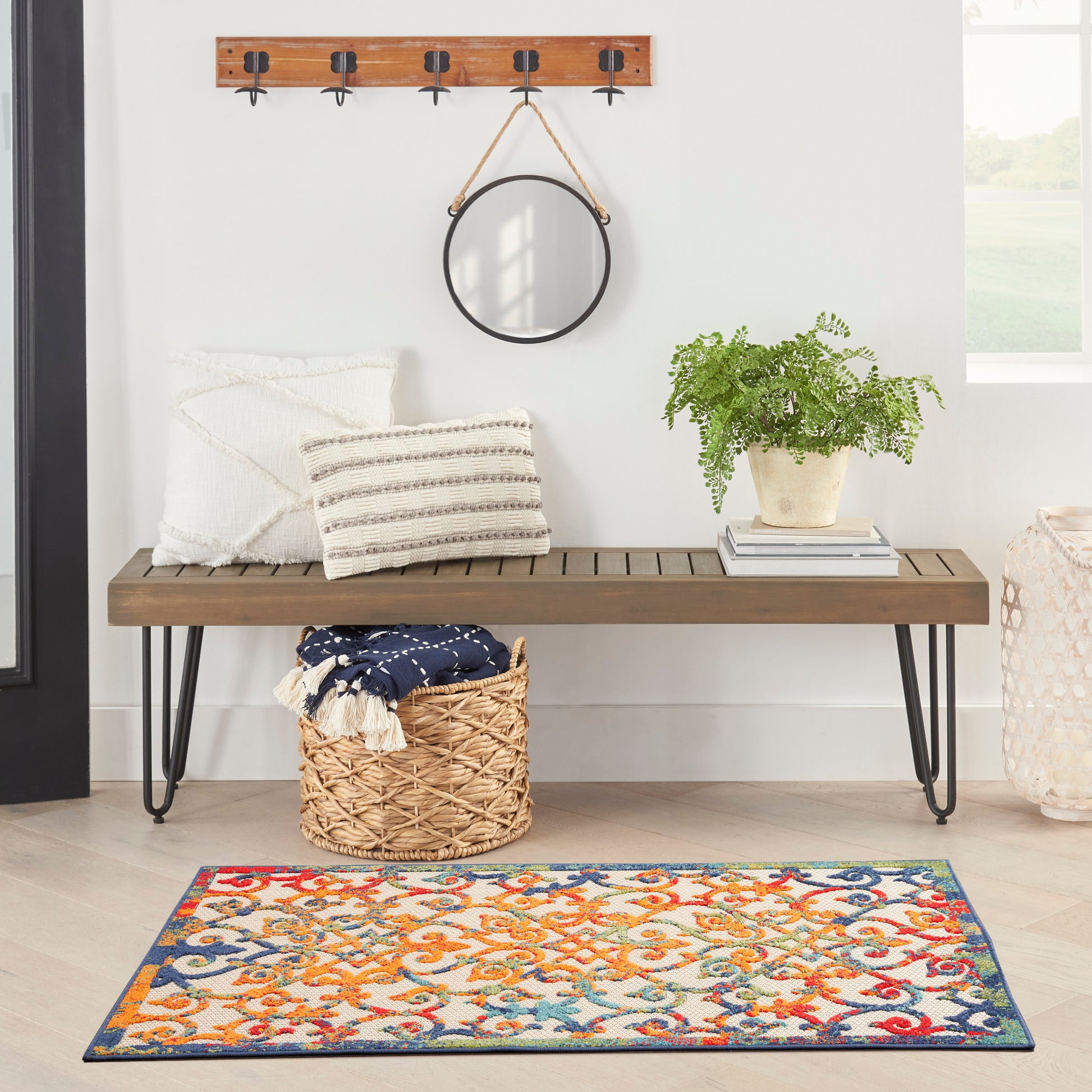 Wood bench with black hairpin legs, striped pillows, round wall mirror, woven basket, potted fern, and vibrant patterned rug.