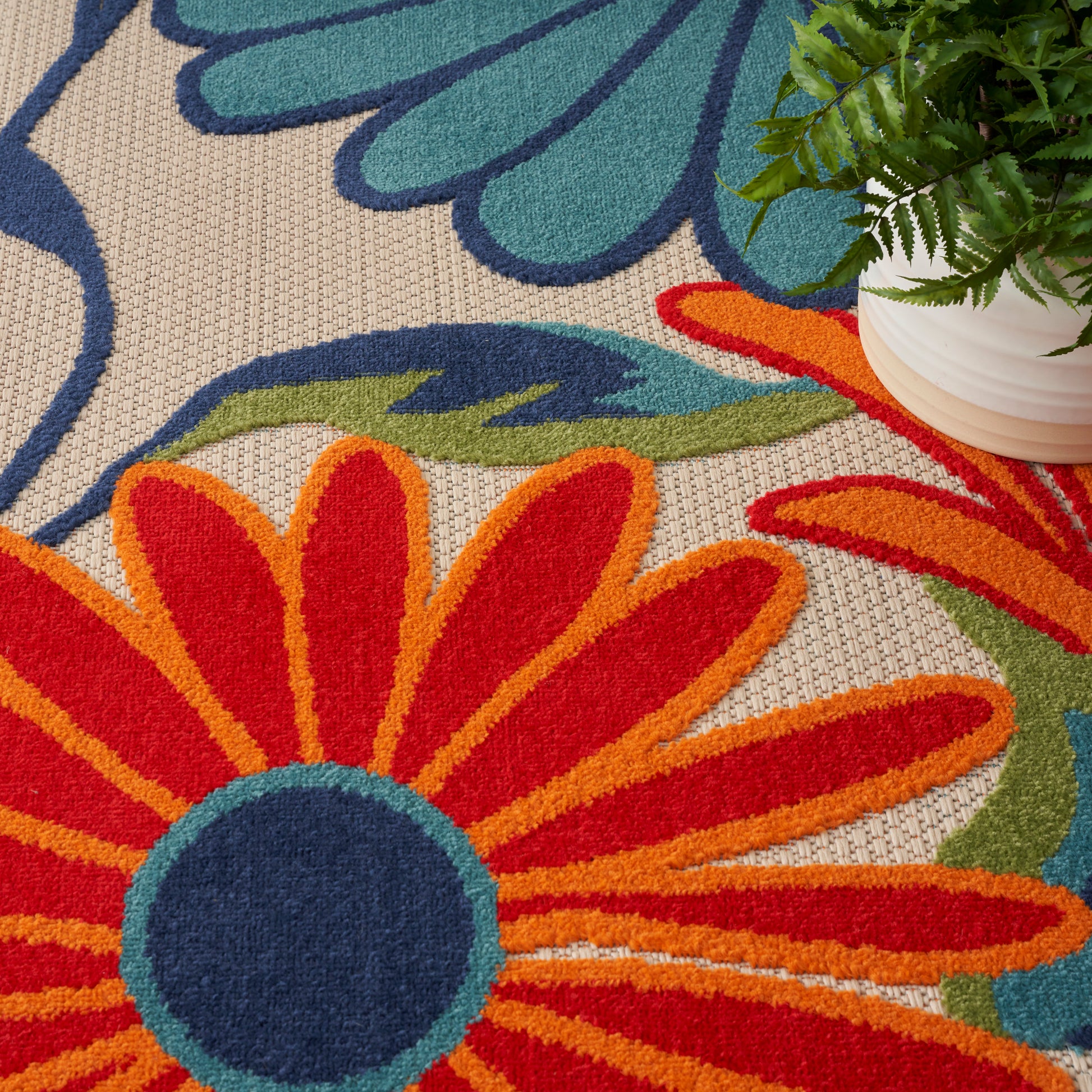 Soft cut-pile rug with bold floral design: orange-red petals, navy center, teal accents on beige background; nearby plant.