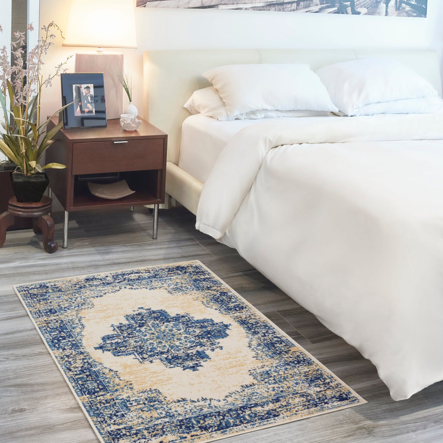 Blue and ivory distressed oriental-style area rug with central medallion on gray wood floor beside a white bed and nightstand.