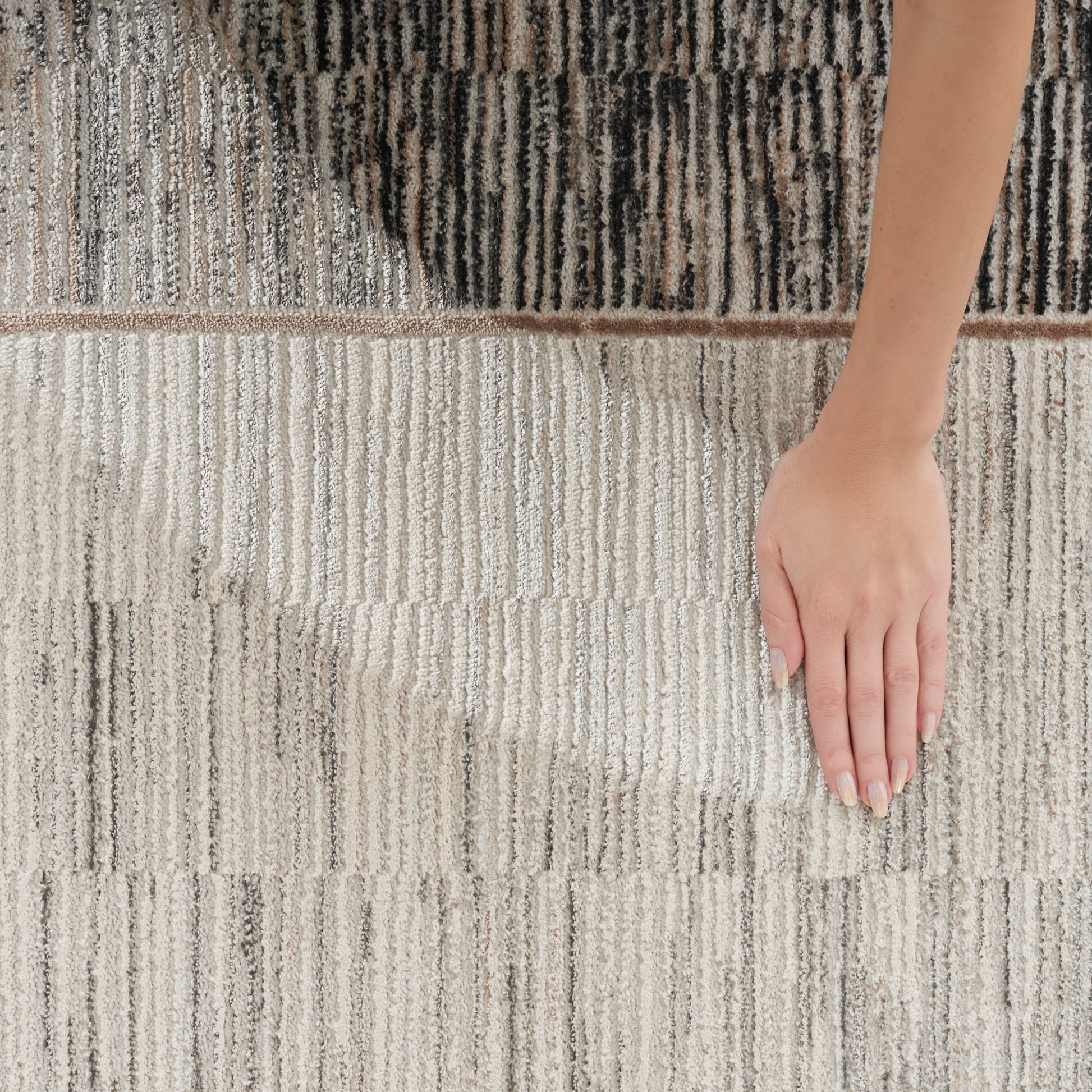 Neutral striped rug with beige, gray and black tones; plush pile, hand resting on surface.