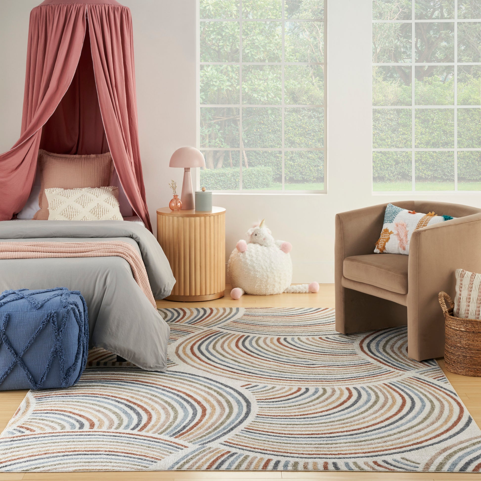 Cozy bedroom with a pink canopy bed, grey bedding, pink lamp, beige armchair, circular patterned rug, large windows.