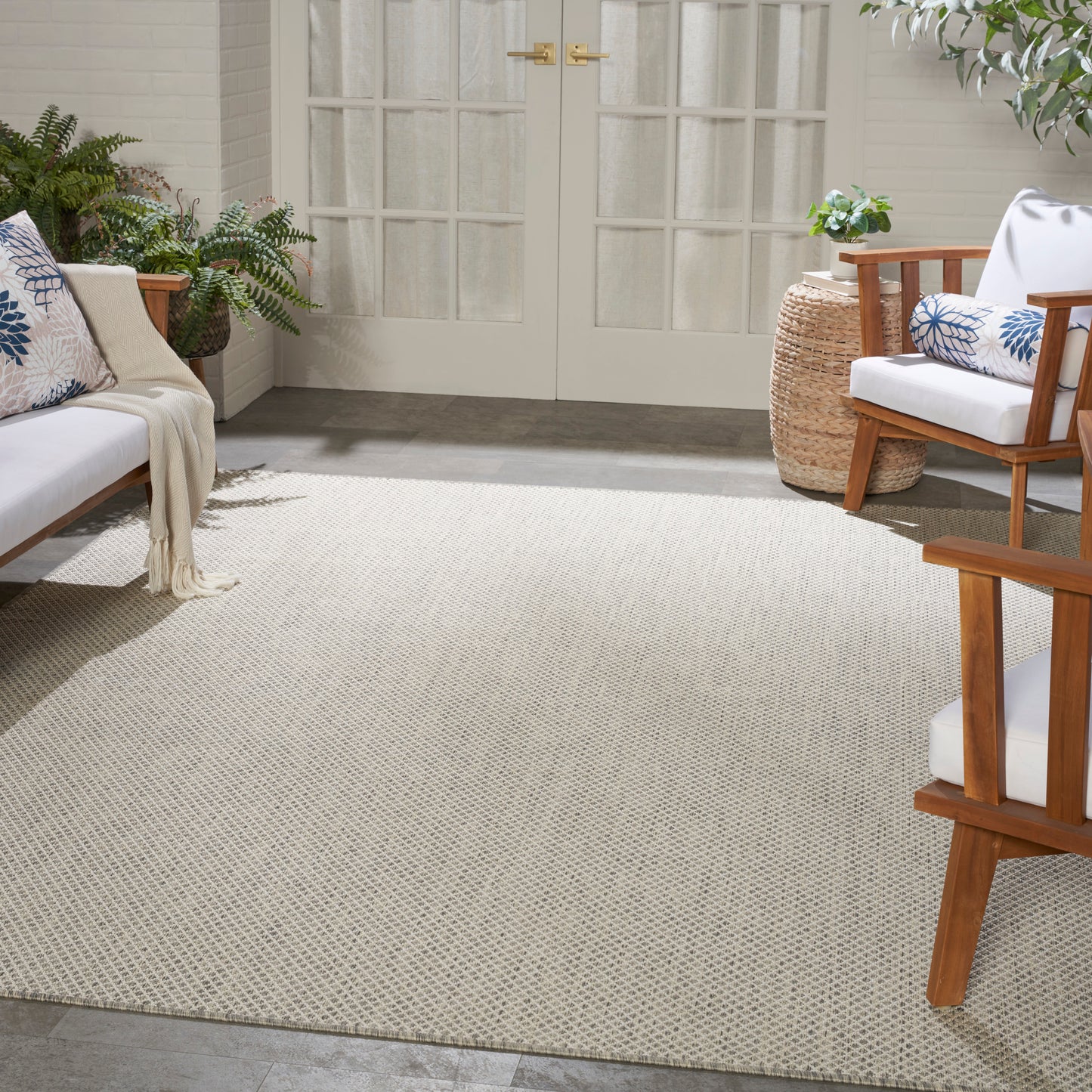 Neutral outdoor living space with beige textured rug, white-cushioned wooden chairs, blue patterned pillows, potted plants & French doors.