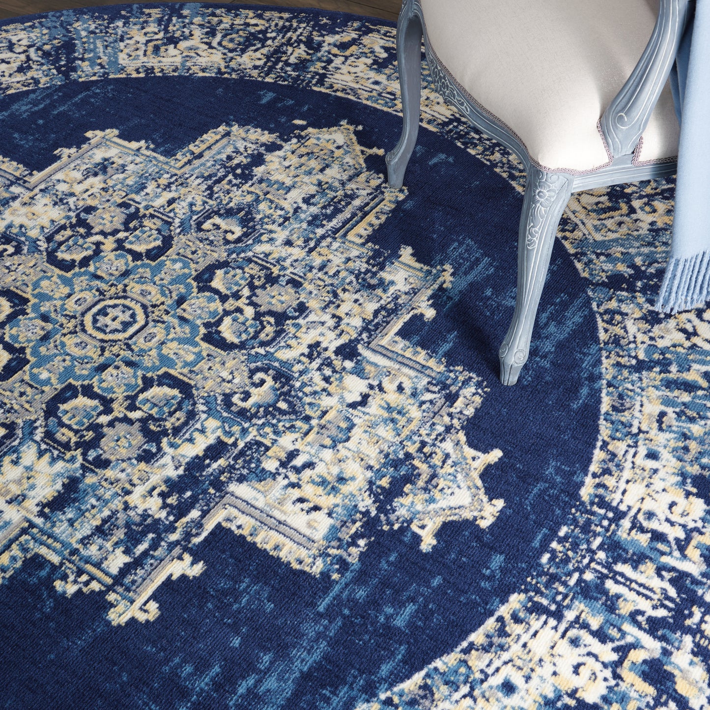 Dark blue oriental rug with ivory and light blue motifs; carved gray-blue chair leg beside beige upholstered seat.