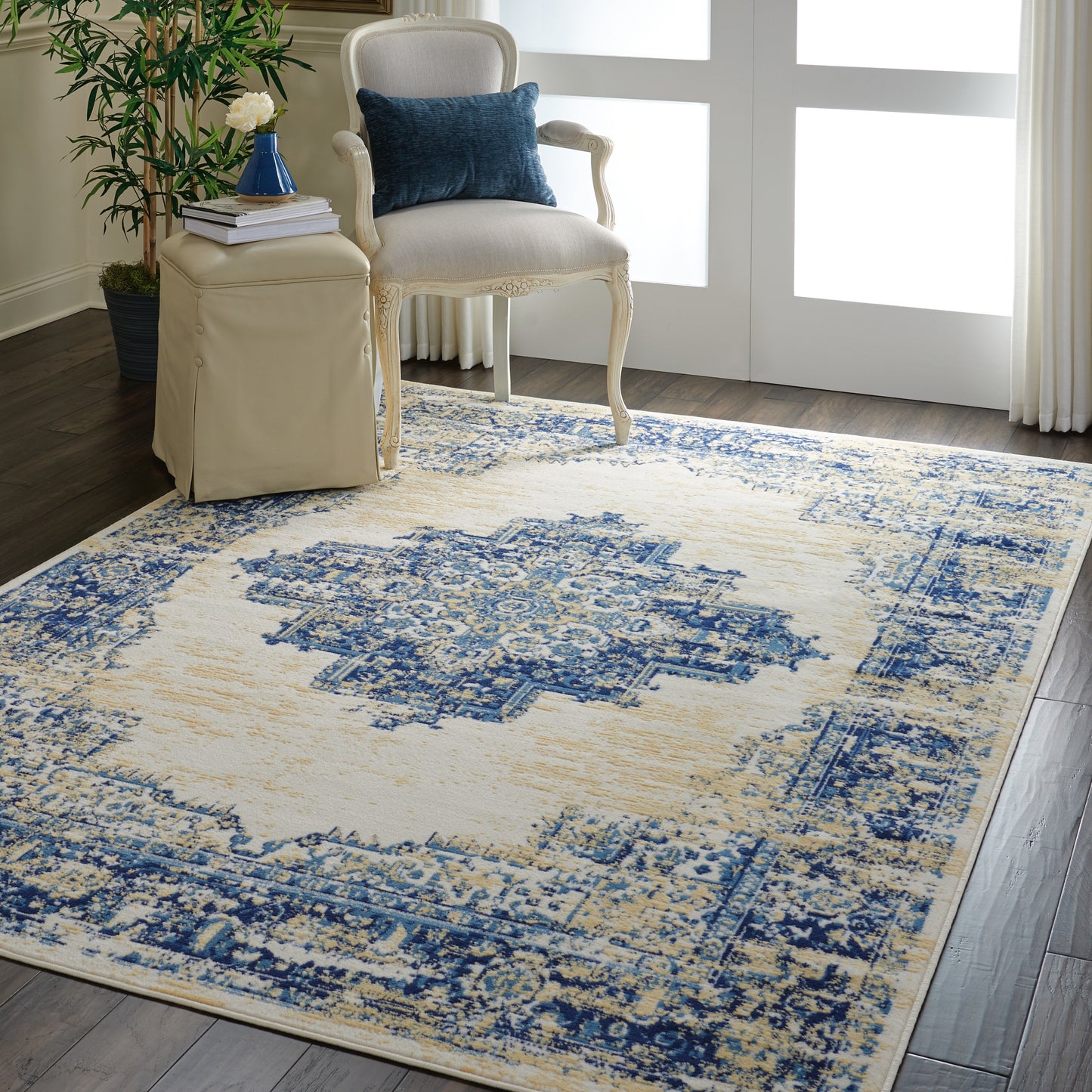 Beige and blue distressed oriental rug in a living room with cream carved-wood armchair, blue pillow, beige ottoman, and potted plant.