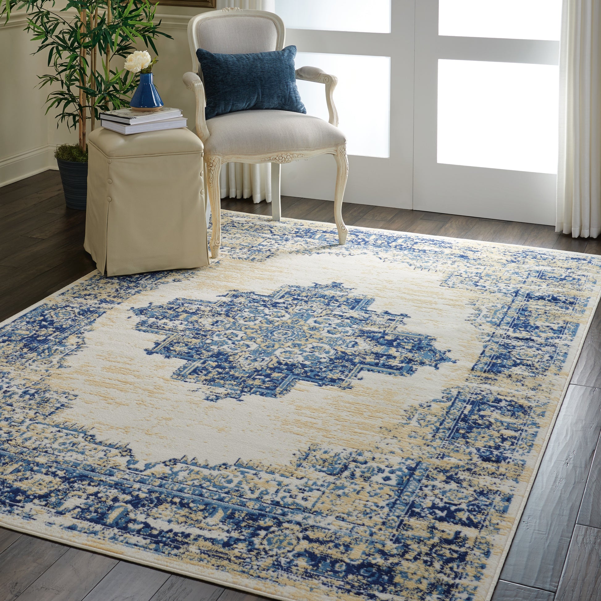 Beige and blue distressed oriental rug in a living room with cream carved-wood armchair, blue pillow, beige ottoman, and potted plant.