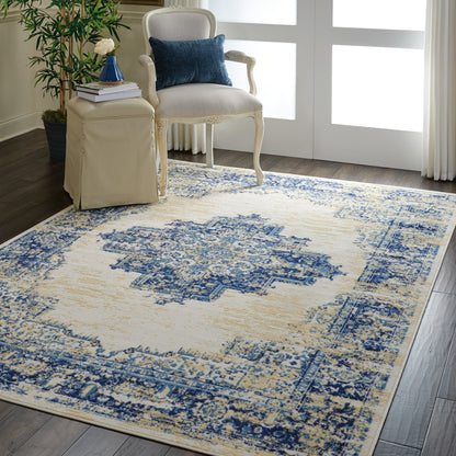 Beige and blue distressed oriental rug in a living room with cream carved-wood armchair, blue pillow, beige ottoman, and potted plant.