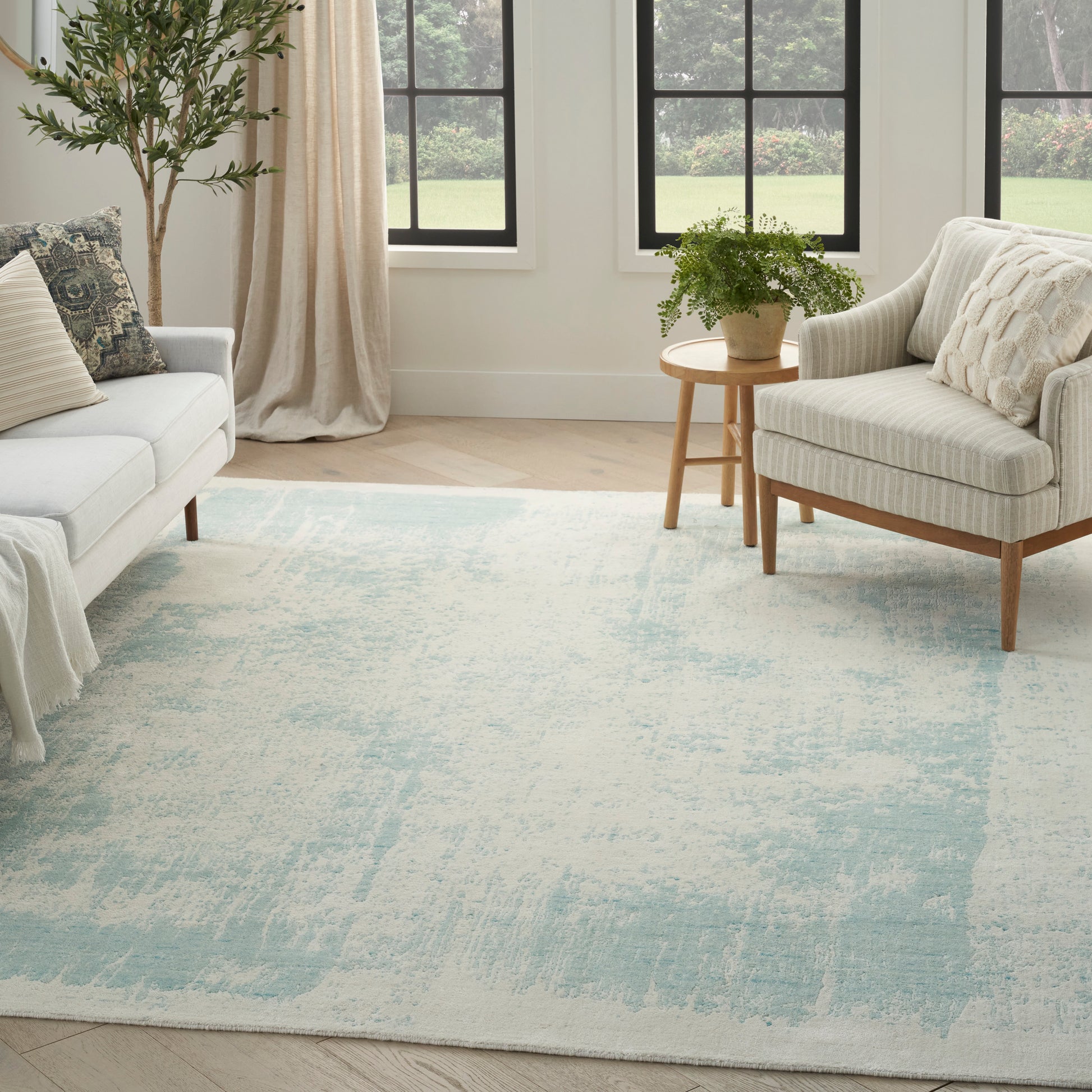 Living room with pale blue/cream rug, beige sofa and armchairs, round wooden side table with plant, beige curtains.