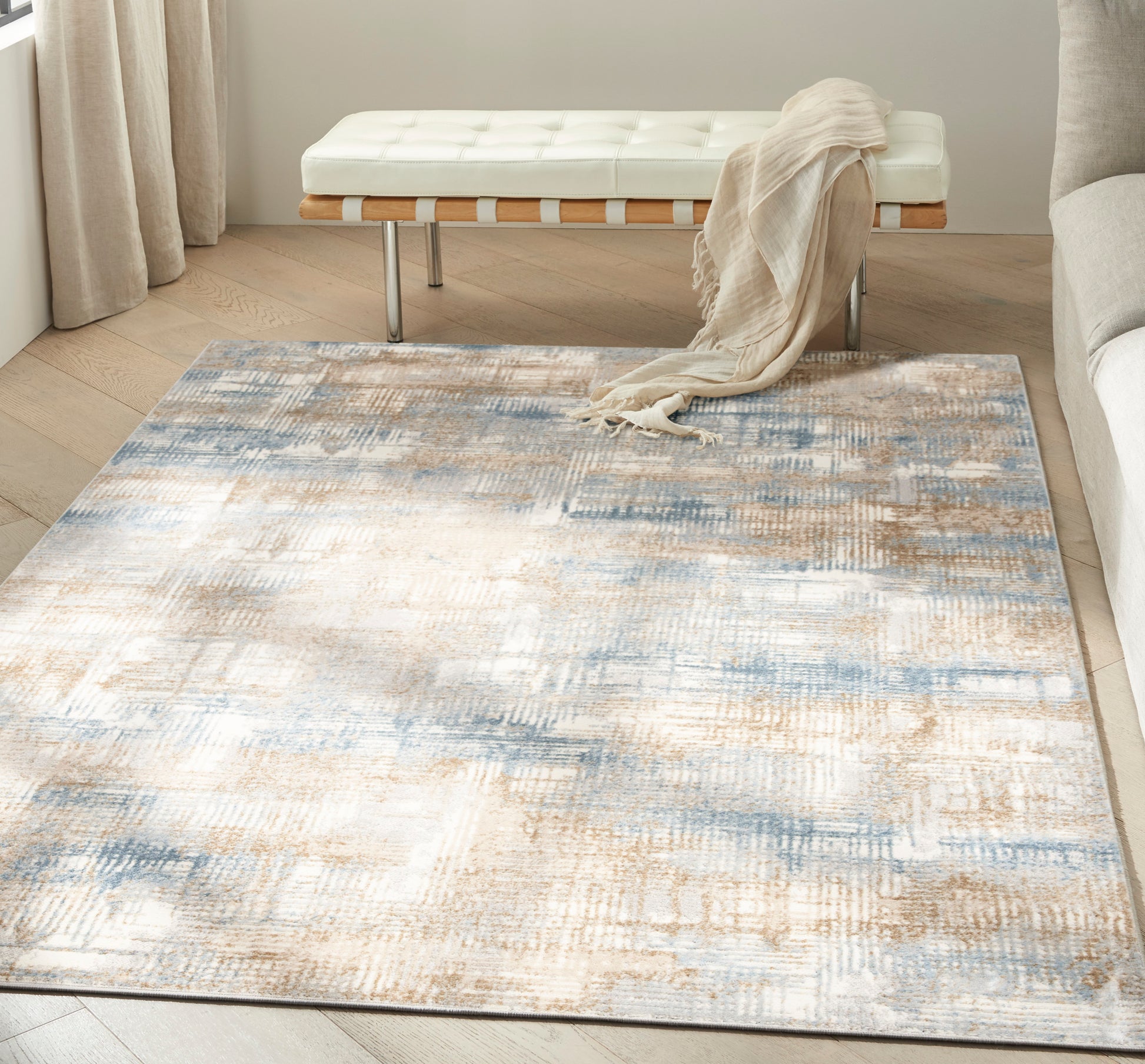 Abstract beige, blue & ivory area rug on light hardwood floor; white tufted bench with wood slats & metal legs, beige throw.