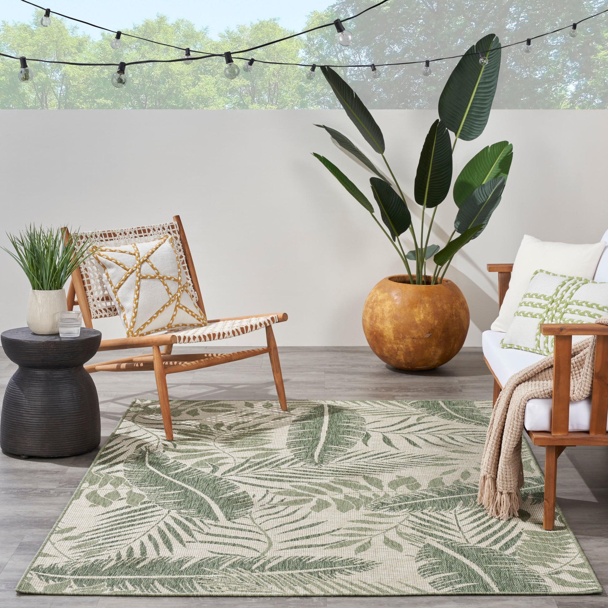 Outdoor living area with palm-leaf rug, wicker chair, white sofa, large potted plant, and string lights overhead.