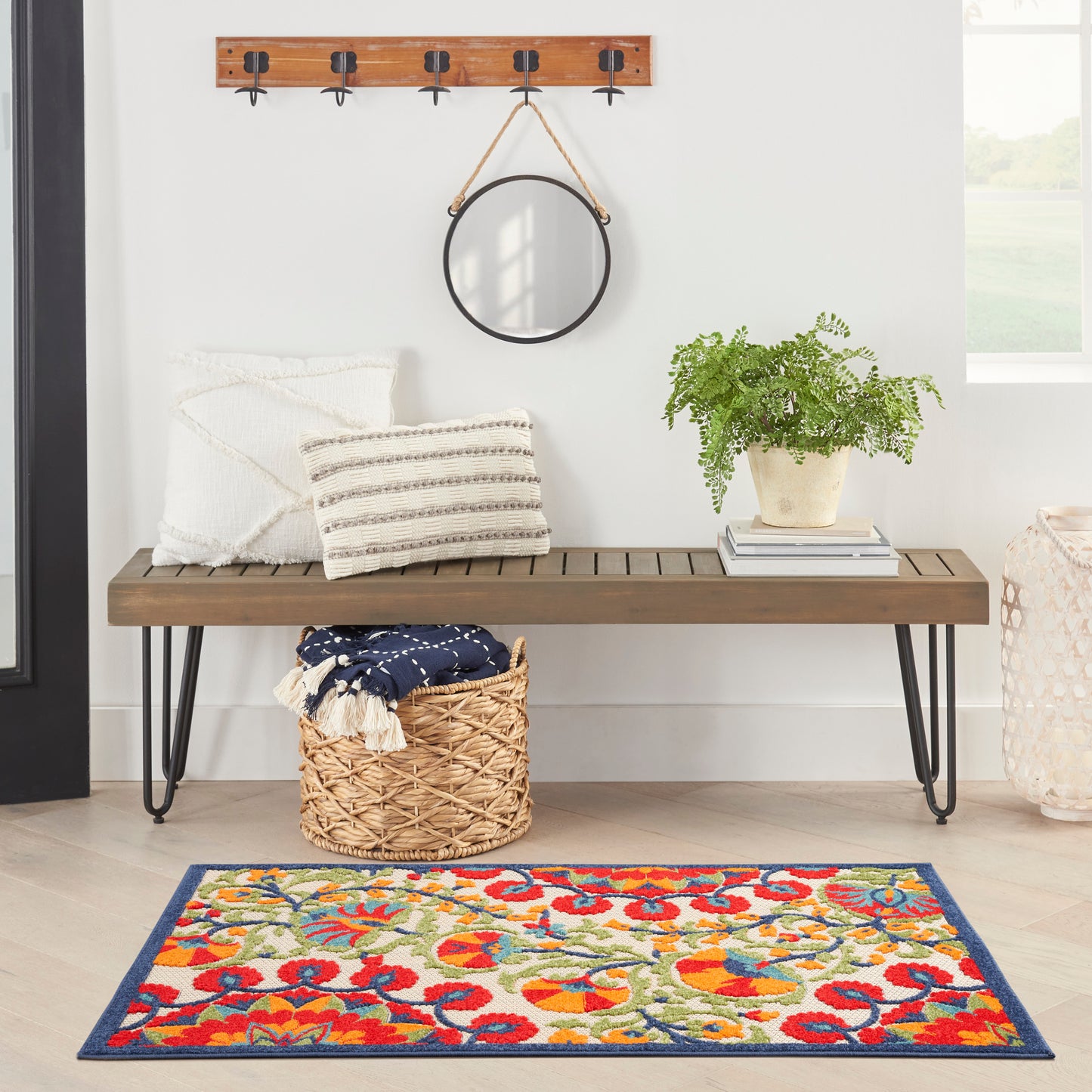 Wood bench with black hairpin legs, white cushions, round wall mirror, potted plant, woven basket, & a colorful rug.