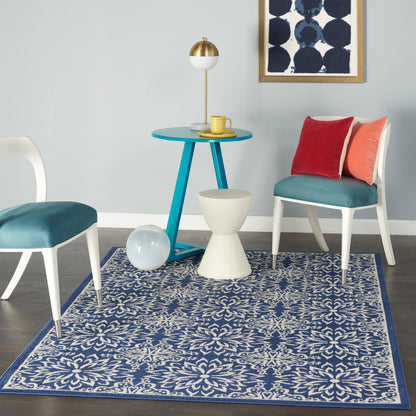 Teal round table with white/teal chairs, red and orange cushions, blue floral rug, gold lamp, and abstract wall art.