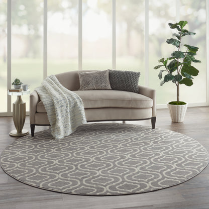 Round grey area rug with white abstract loop pattern in a modern living room; beige sofa, knitted throw, and potted plant.