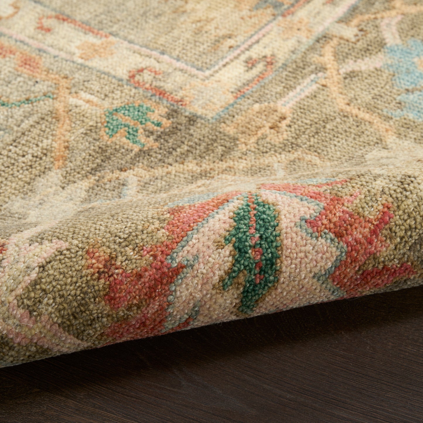 Beige oriental rug with pink, green & teal accents; dense wool pile, folded edge, on dark wooden floor.
