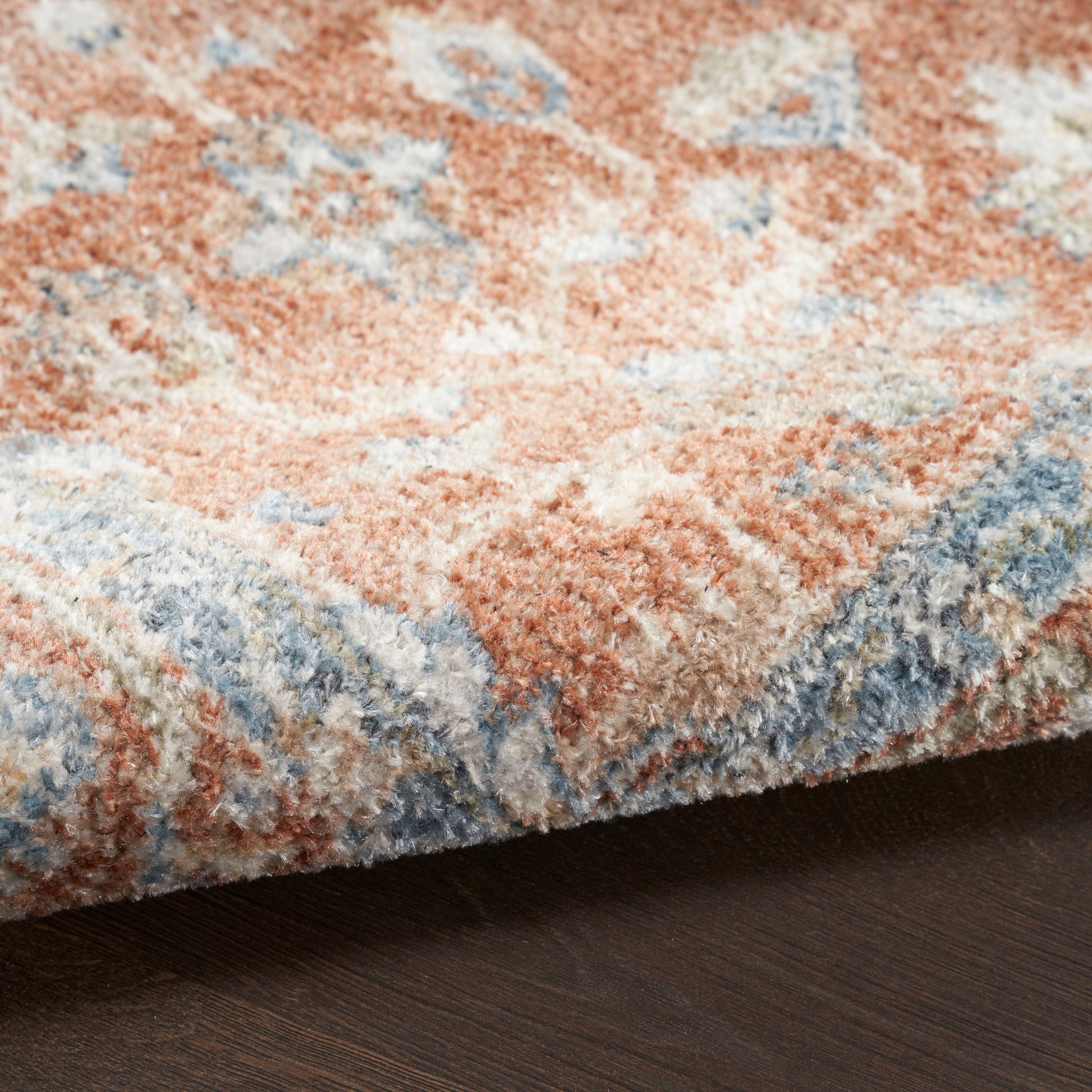 Close-up of a plush patterned area rug in peach, blue and ivory tones, edge folded over a dark wood floor.