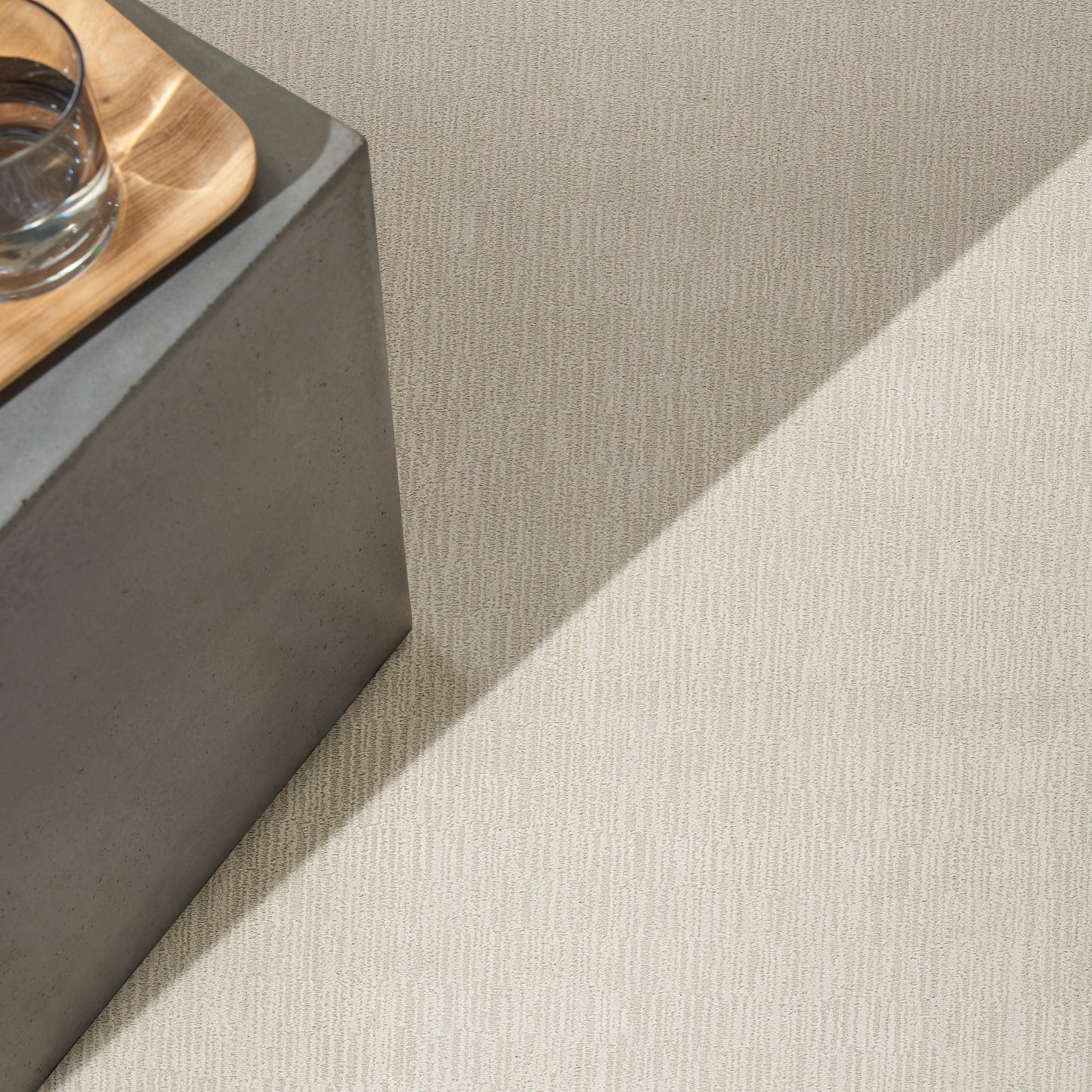 Grey concrete block pedestal with a light wood tray and glass on beige textured carpet; minimalist modern decor.