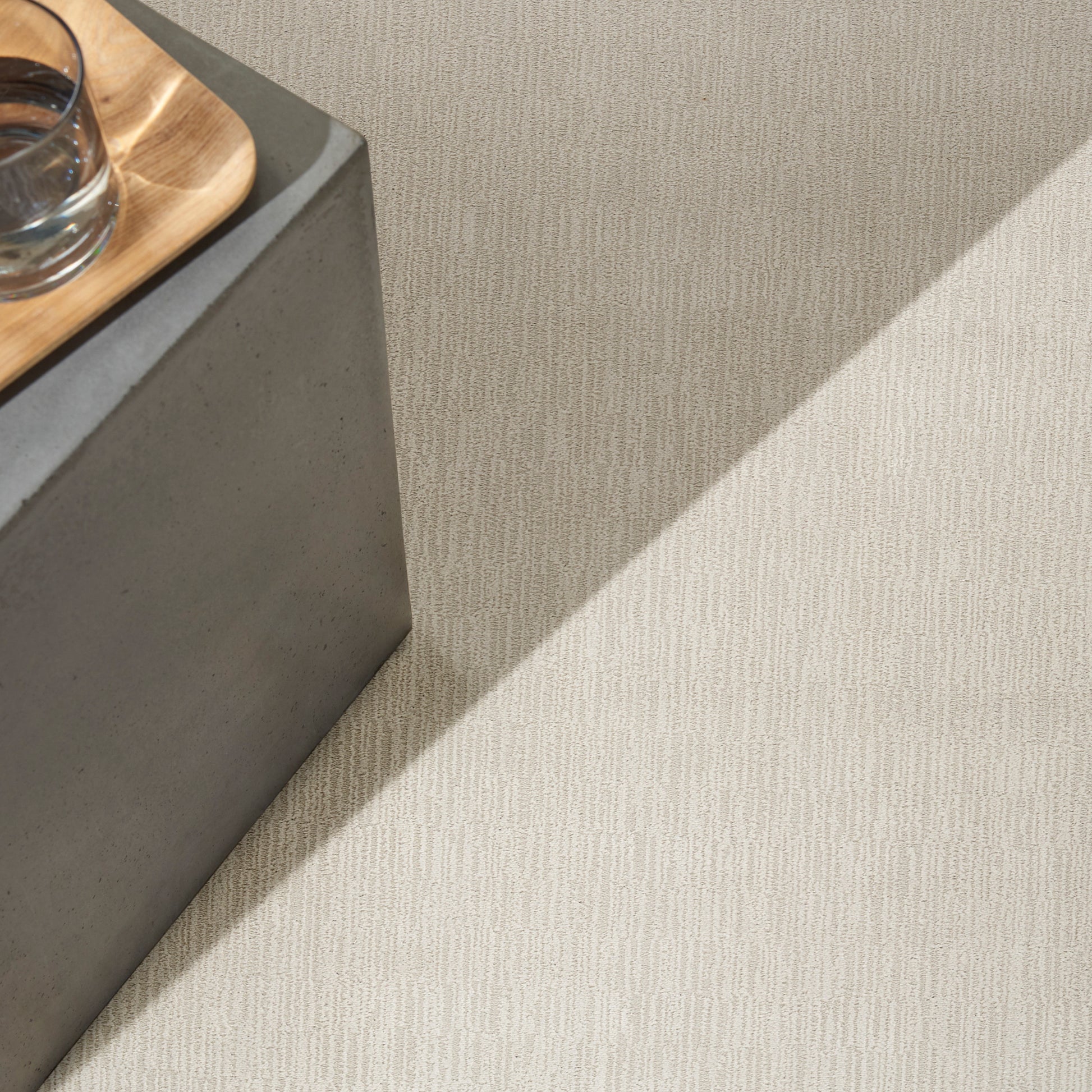 Grey concrete block pedestal with a light wood tray and glass on beige textured carpet; minimalist modern decor.