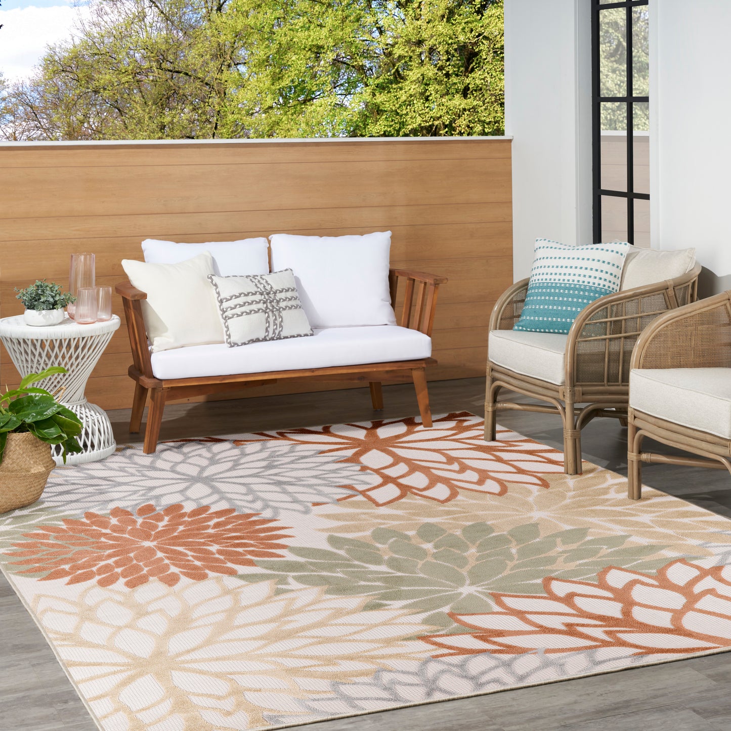 Outdoor lounge area with wooden sofa, rattan chairs, white cushions, and a large floral rug in orange, teal, beige.