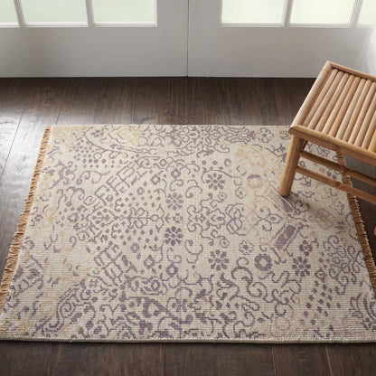 Rectangular beige rug with lilac floral pattern, fringe edges, on dark wood floor; wooden slatted stool sits nearby.