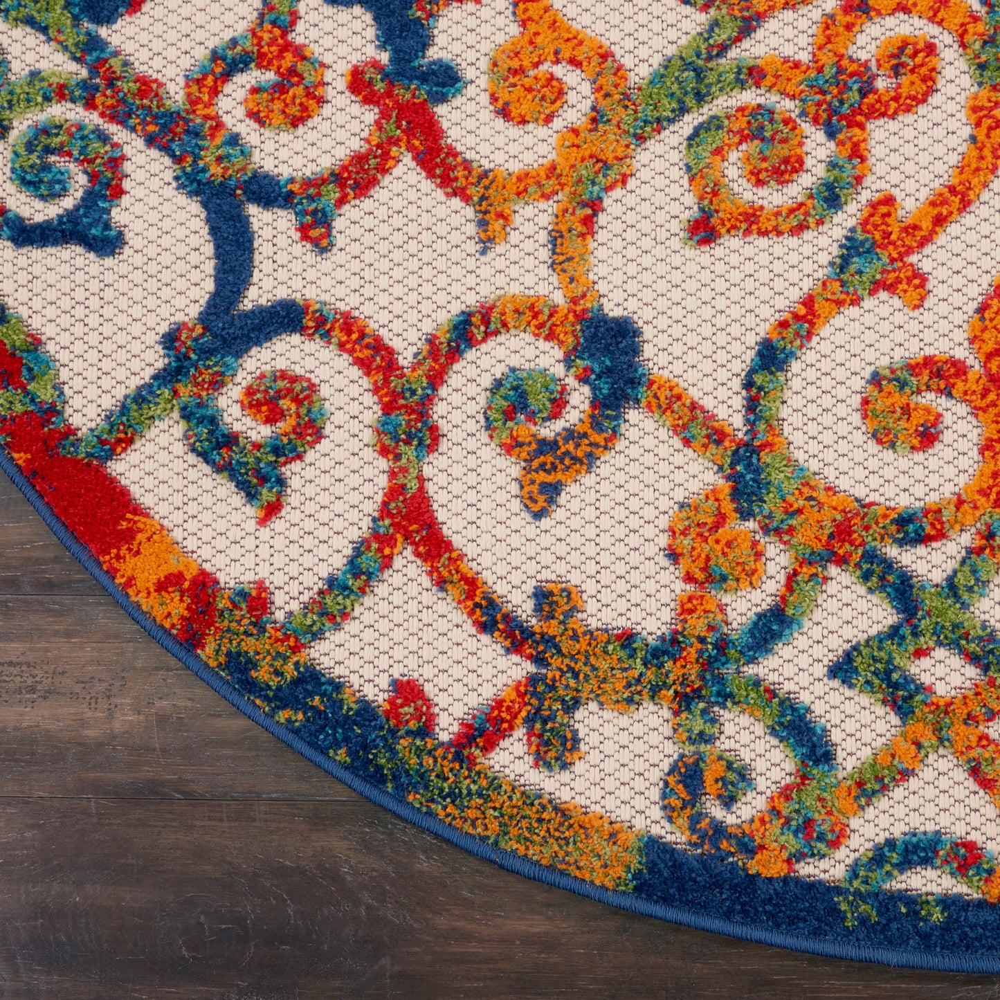 Round tufted rug with ornate scrolling motif in red, orange, blue & green on beige; blue edge binding on dark wood floor.