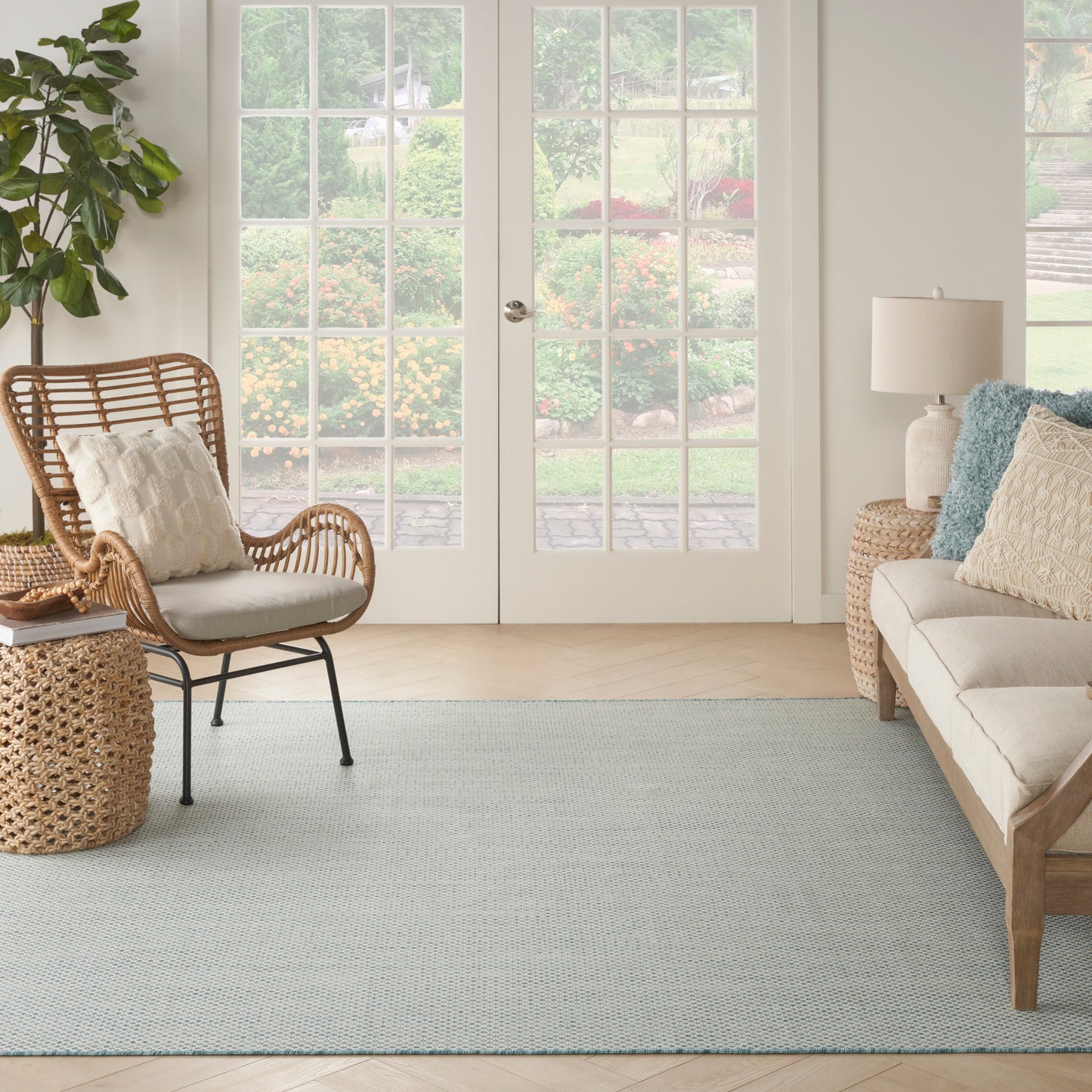 Bright living room with beige sofa, rattan chair, woven side table, pale blue rug, greenery, French doors.