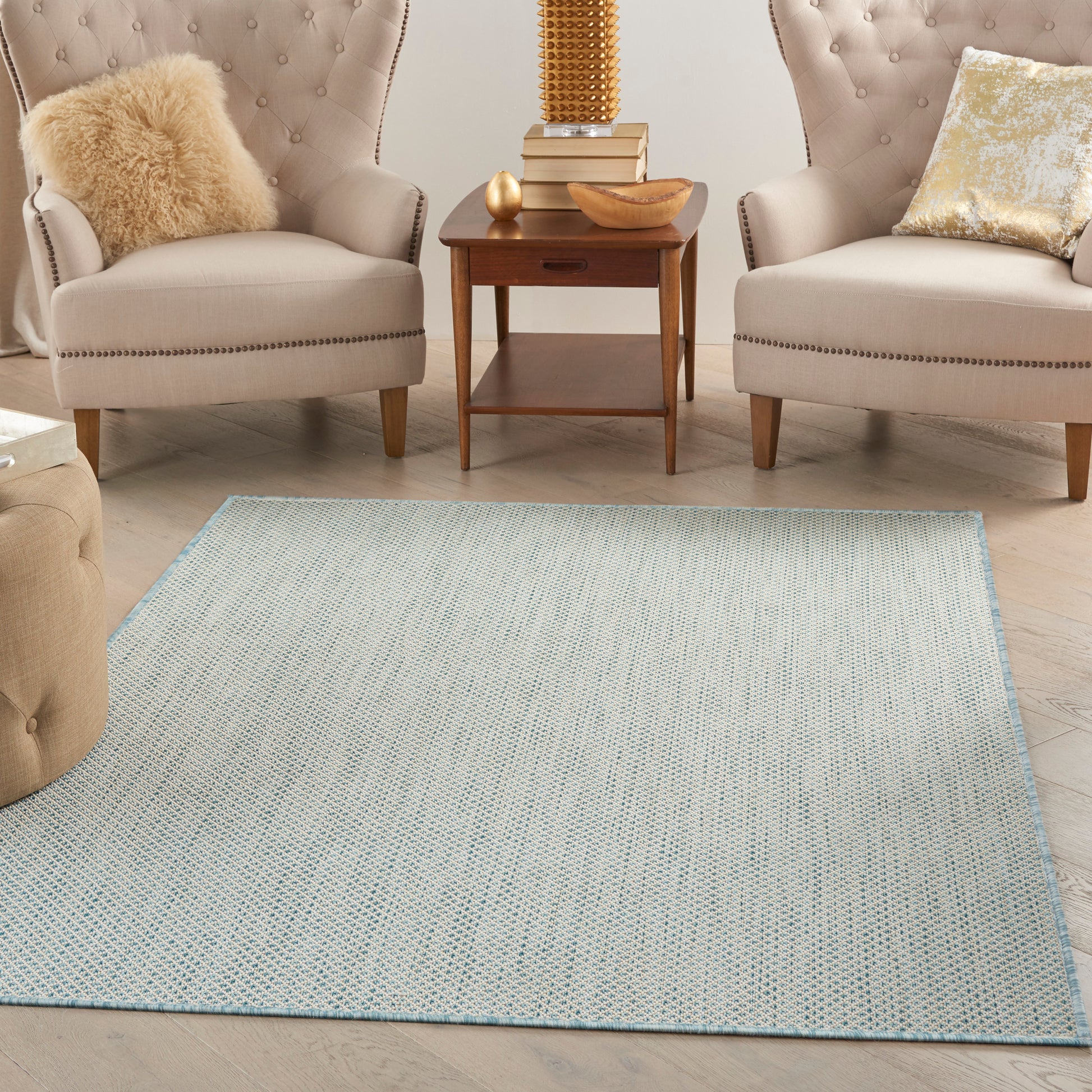 Two beige tufted armchairs with nailhead trim flank a wooden end table on a pale blue woven rug.