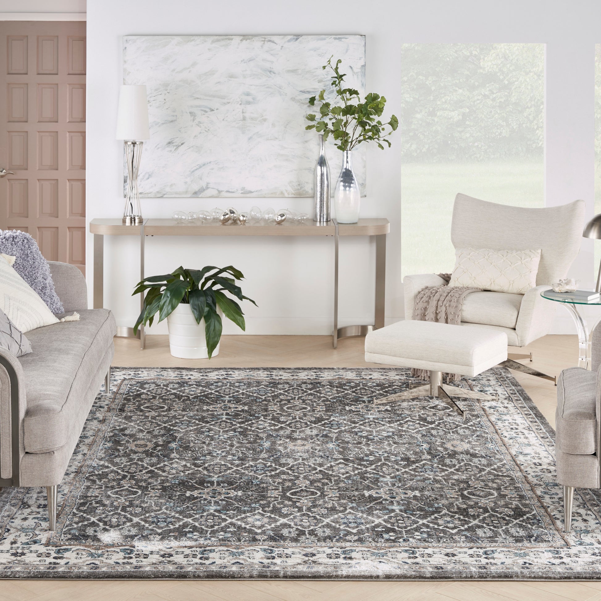 Modern living room with gray upholstered sofas, beige chair, blue-gray patterned rug, plants, and a console table with lamp.