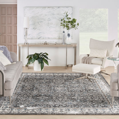 Modern living room with gray upholstered sofas, beige chair, blue-gray patterned rug, plants, and a console table with lamp.