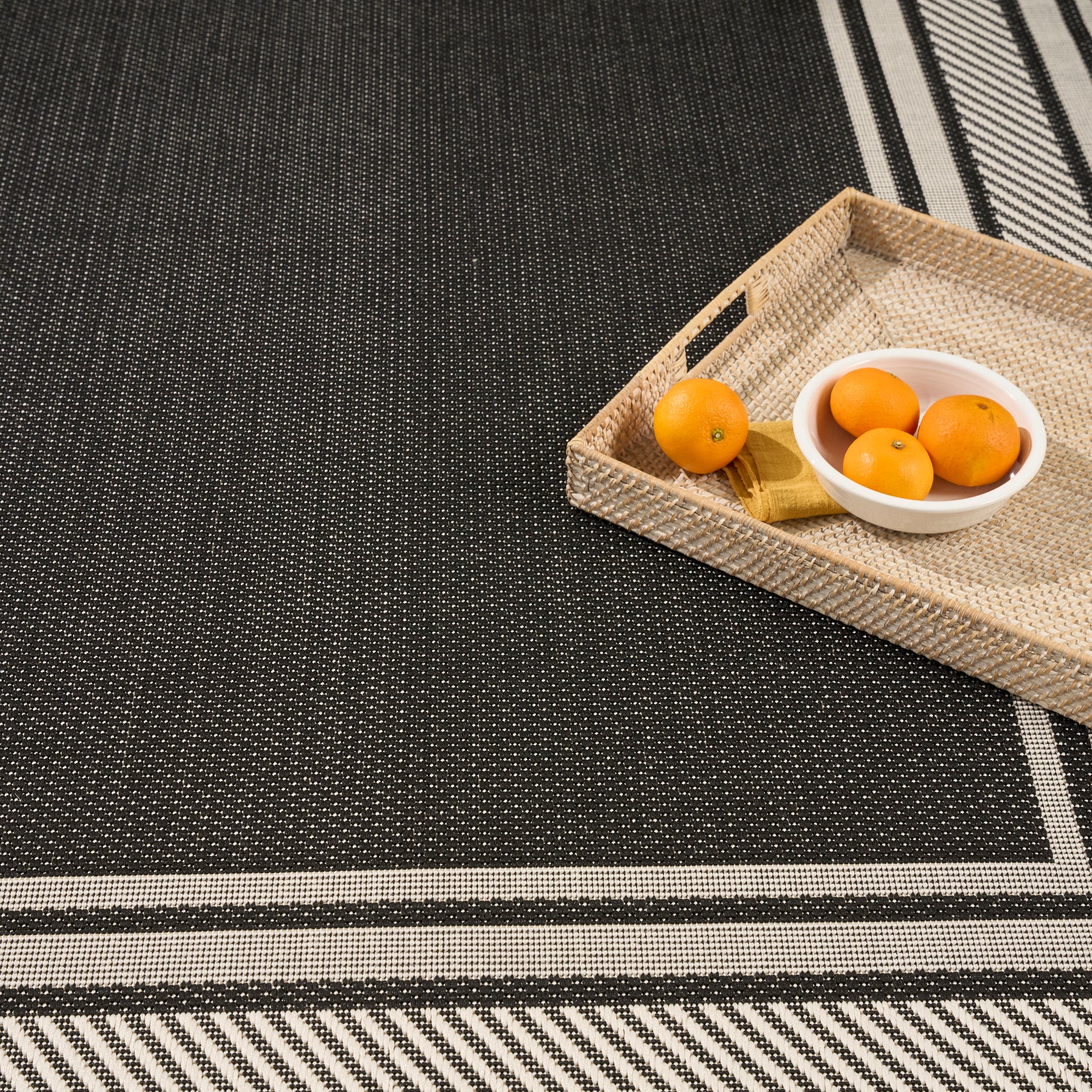 Black textured rug with white border stripes; beige wicker tray with a white bowl of three oranges.