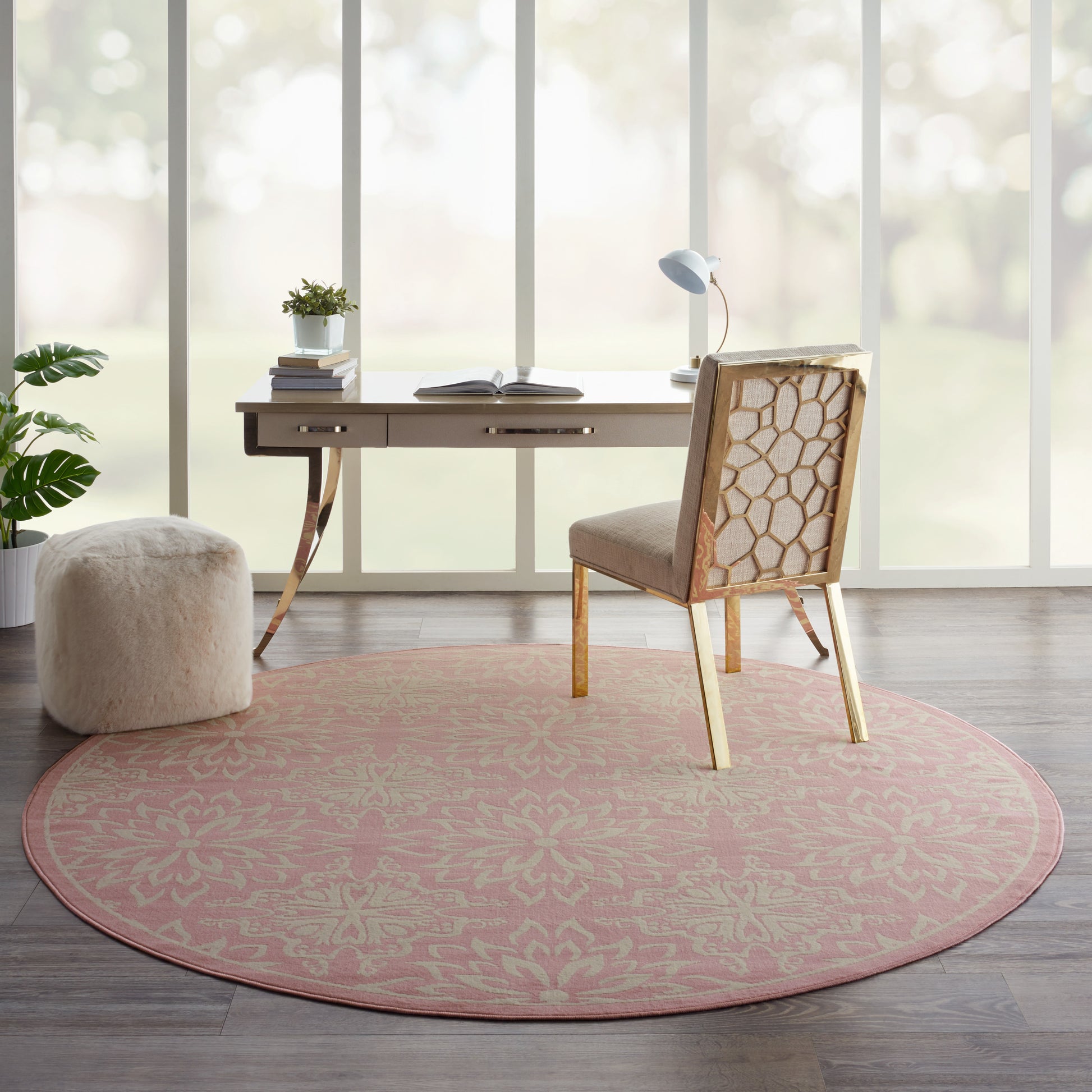 Round pink patterned rug under a light wood desk and lattice-back chair in a bright home office with plant, books, and lamp.