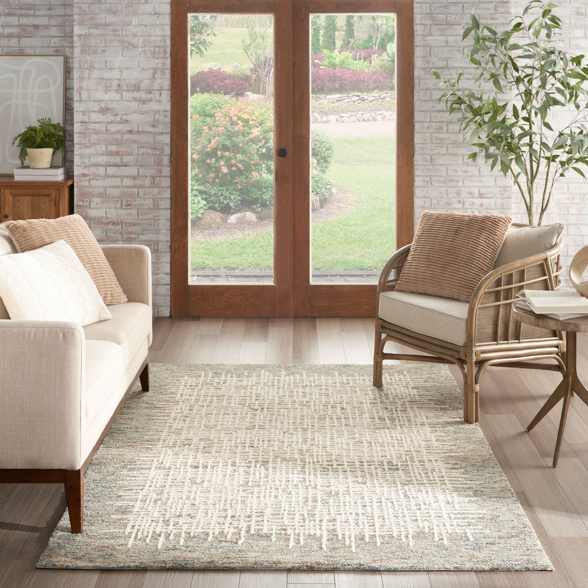 Cozy living room with beige sofa, rattan armchair, neutral patterned rug, brick wall, and wood-framed glass doors to a garden.