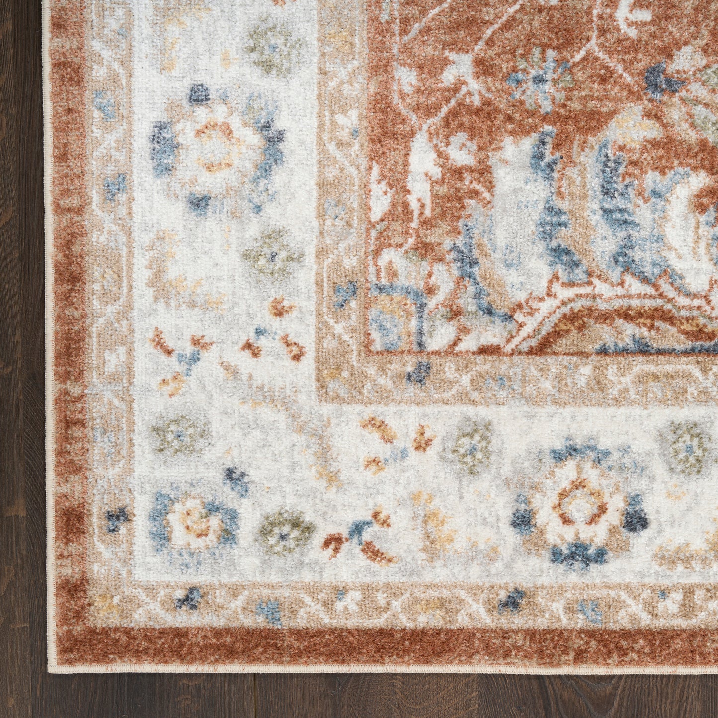 Close-up of ornate area rug corner with ivory border, rust center and blue floral accents on dark wood floor.