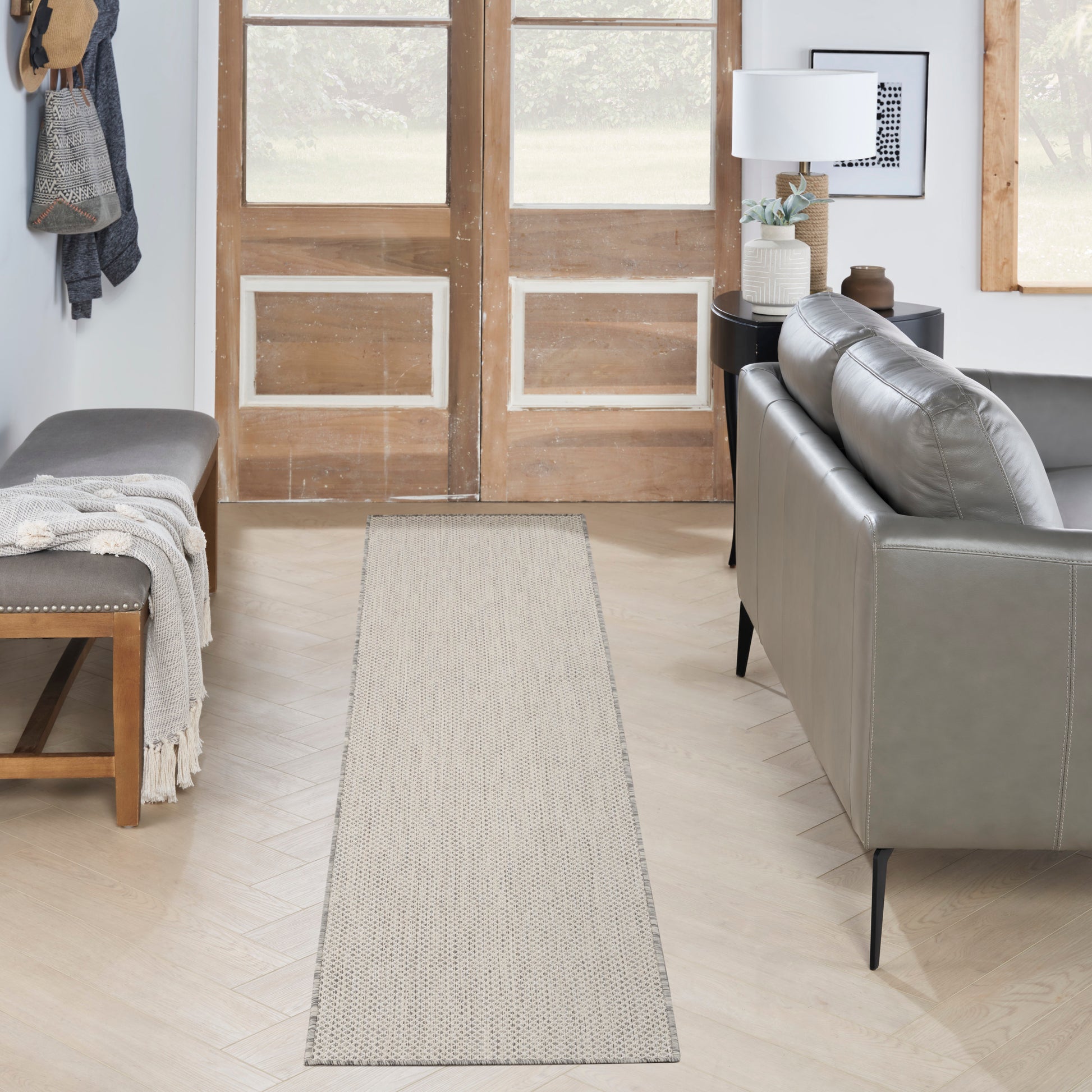 Neutral entryway with long beige rug runner, light wood floor, gray leather sofa on the right, bench with blanket on left.