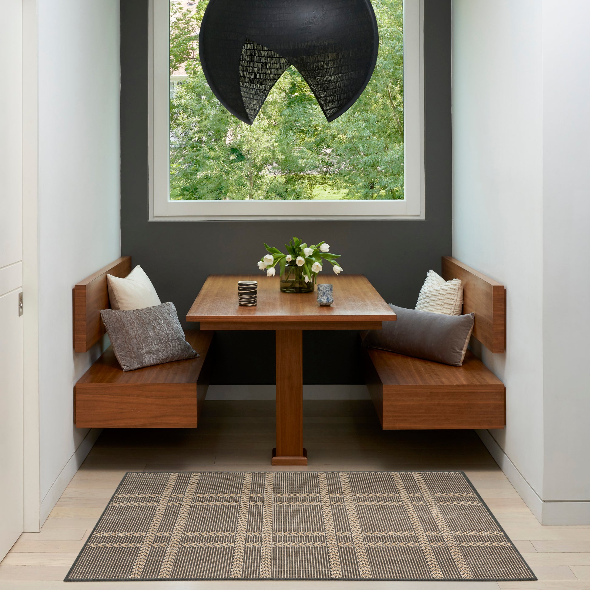 Wood banquet nook with L-shaped benches, table, grey cushions, vase of white flowers, black curved pendant lamp, plaid rug.