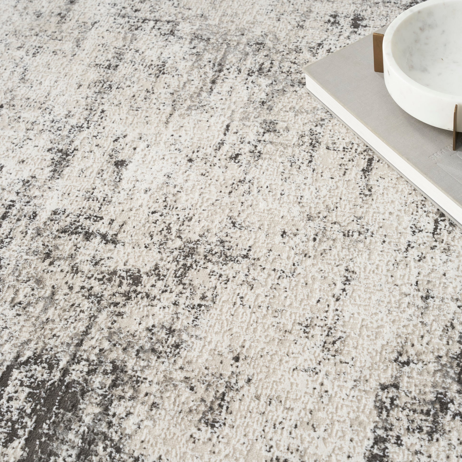 Beige and gray abstract-pattern rug with textured fibers, plus a white marble bowl and neutral-toned books in the top-right.