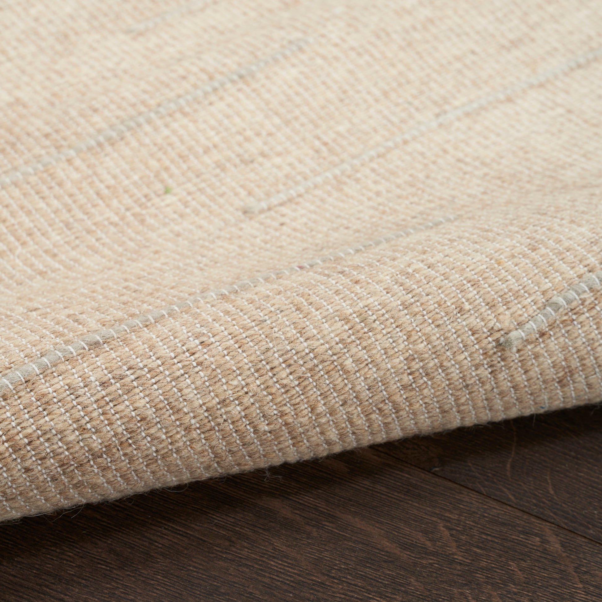 Beige flatweave rug in natural fibers with subtle stripe pattern, folded edge on dark hardwood floor.