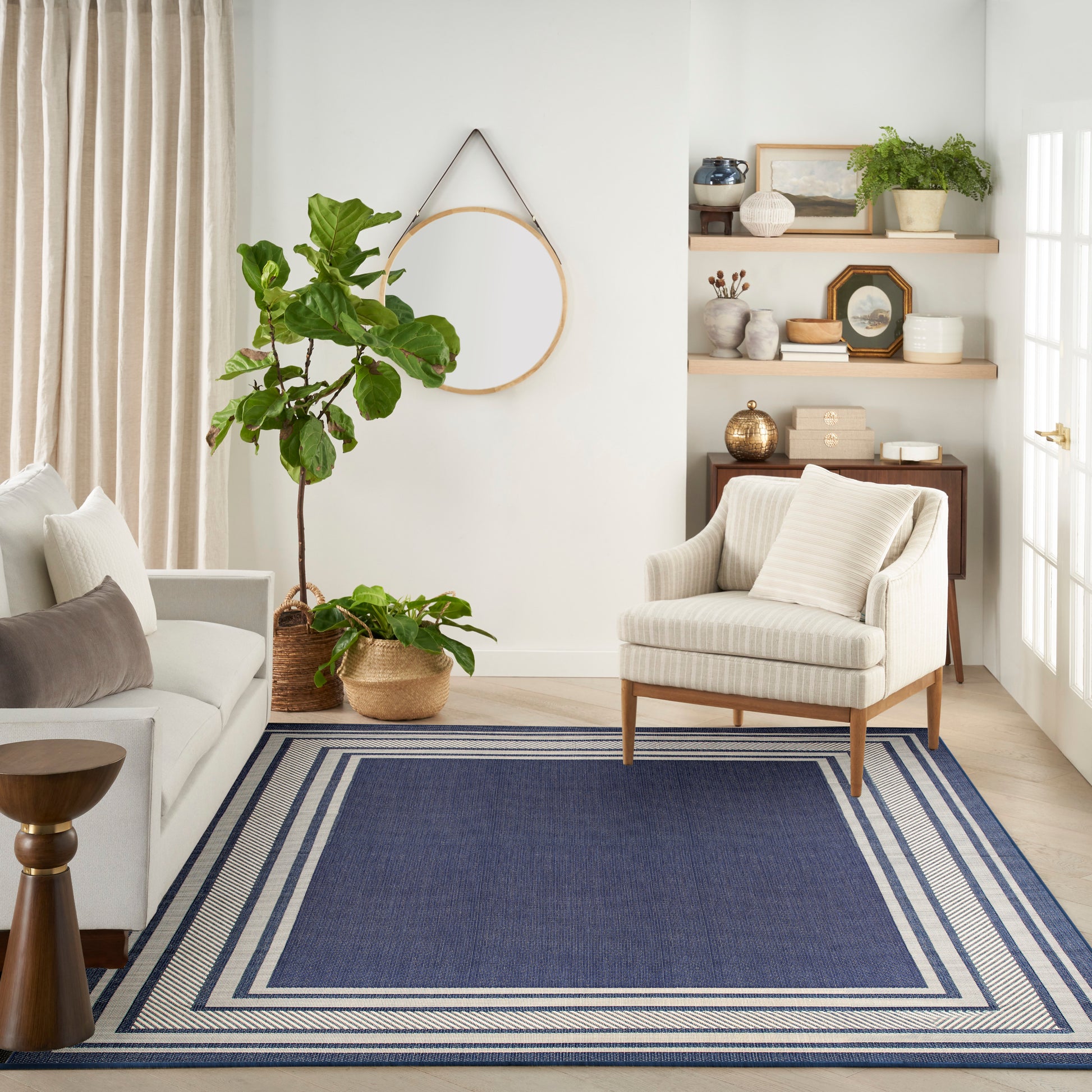 Bright living room with blue bordered rug, cream sofa and armchair, tall indoor plant, hoop decor and wooden shelves.