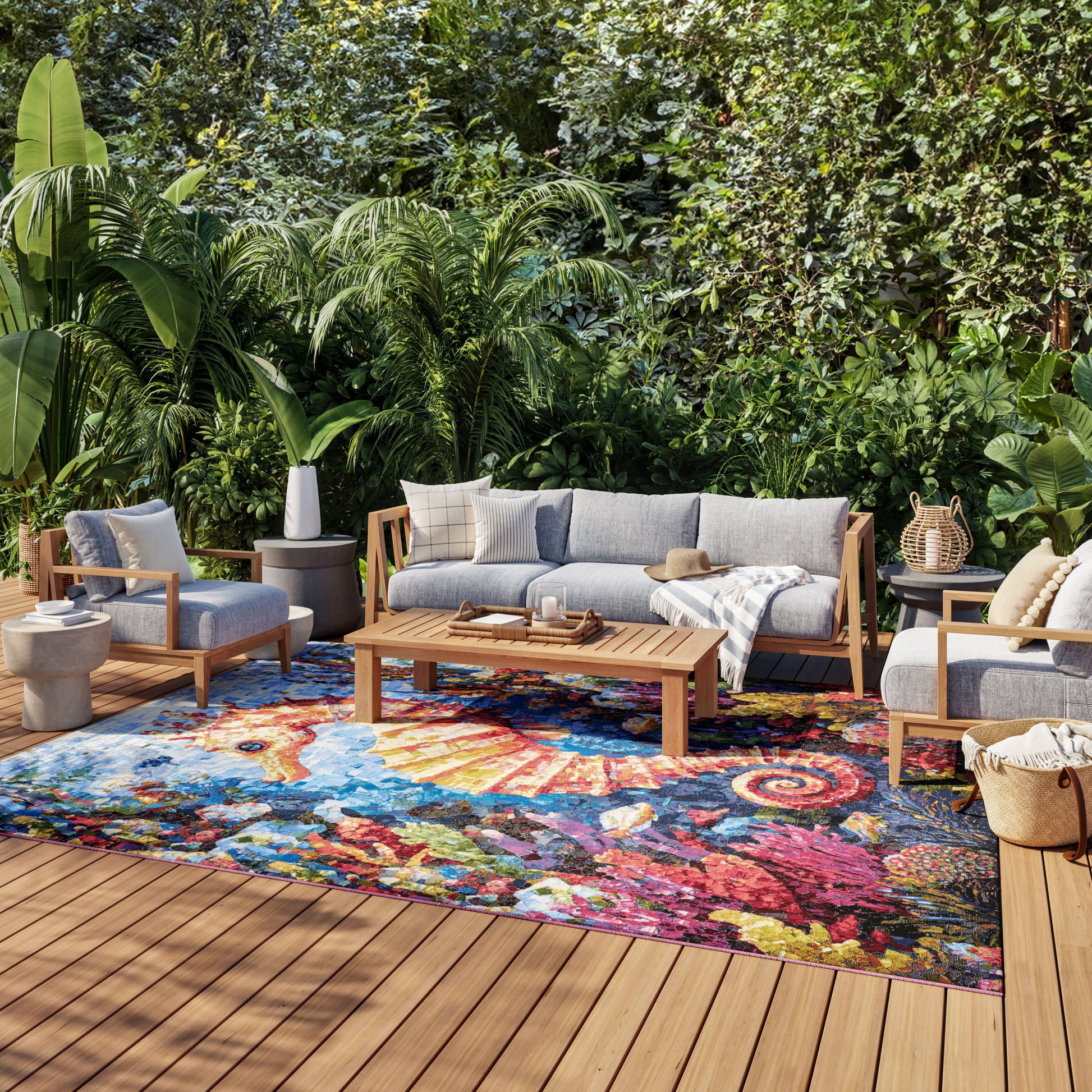 Outdoor lounge with gray-cushion wooden sofa and chairs on a colorful abstract rug amid lush tropical plants.