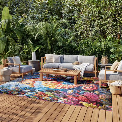 Outdoor lounge with gray-cushion wooden sofa and chairs on a colorful abstract rug amid lush tropical plants.