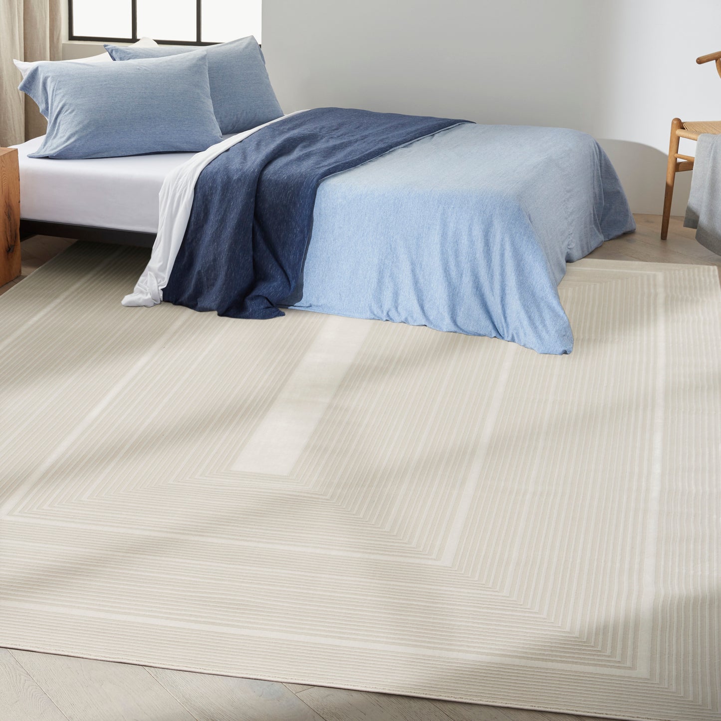 Bed with light-to-navy blue bedding on a beige geometric rug; airy room, window light, wooden chair nearby.