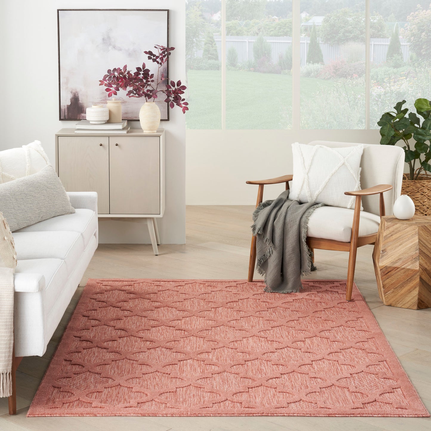 Living room with a coral-pink patterned rug, white sofa, wooden armchair, gray throw and light wood furniture.