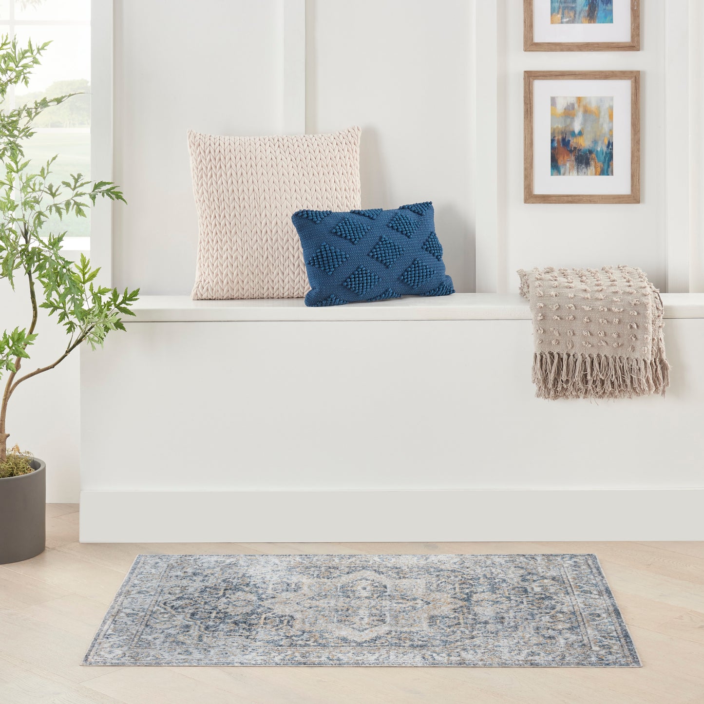 Beige knitted square cushion, navy blue diamond-textured cushion, beige fringe throw on a white bench; blue rug, plant, art.