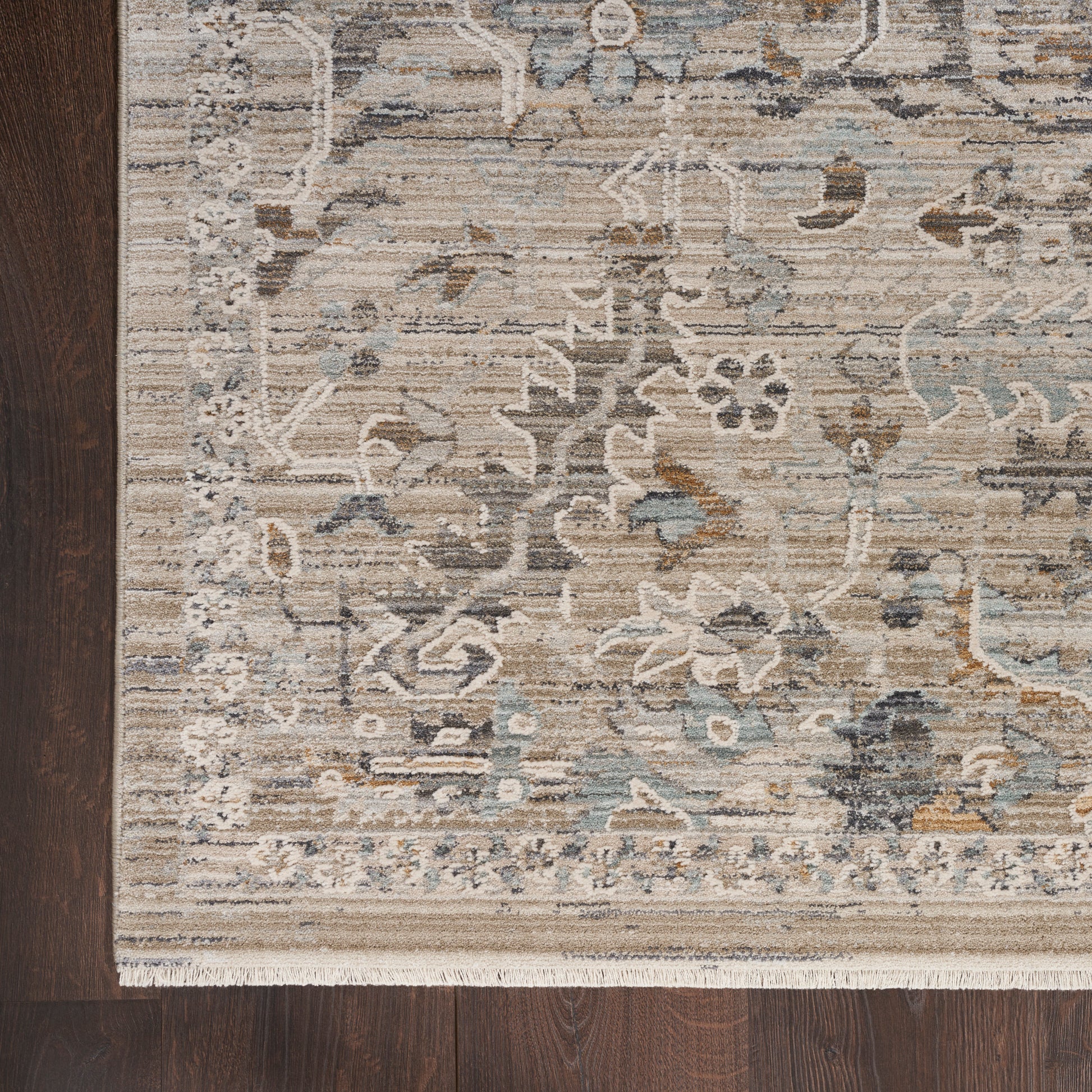 Beige oriental rug with blue-gray floral pattern, cream fringe along bottom, placed on dark wood floor.