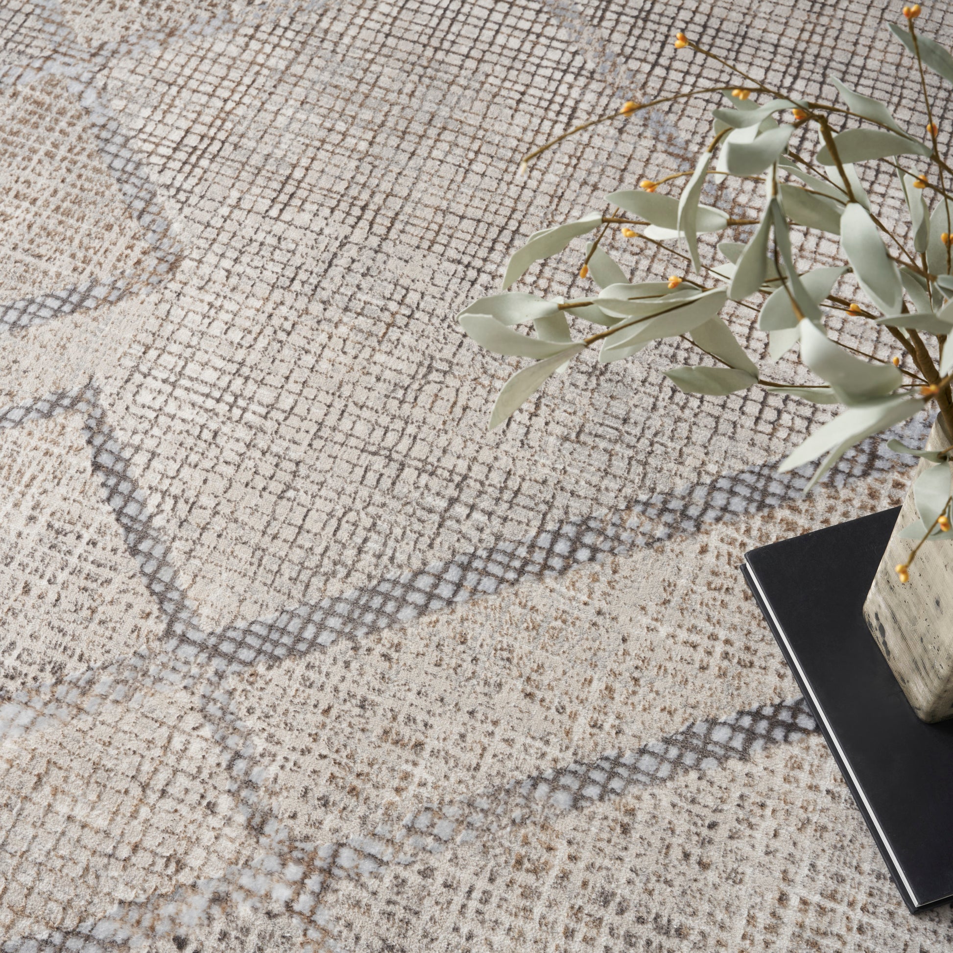 Beige textured rug with gray diamond lattice pattern; ceramic vase with eucalyptus leaves beside a black notebook.
